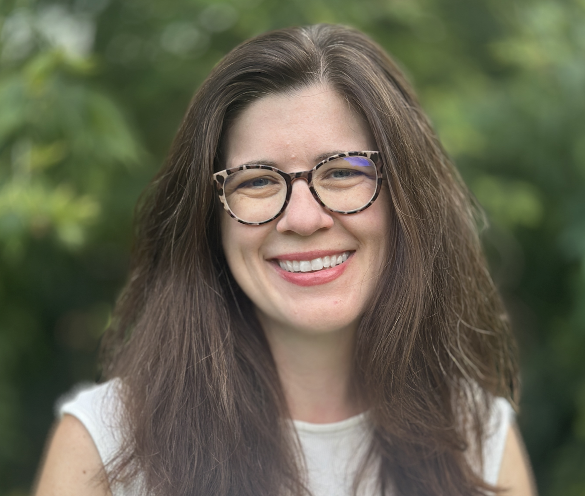 A woman with long brown hair, wearing tortoiseshell glasses, smiling outdoors with green foliage in the background.