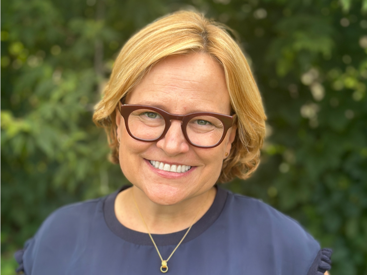 A woman with short blonde hair and glasses smiling outdoors with green foliage in the background.