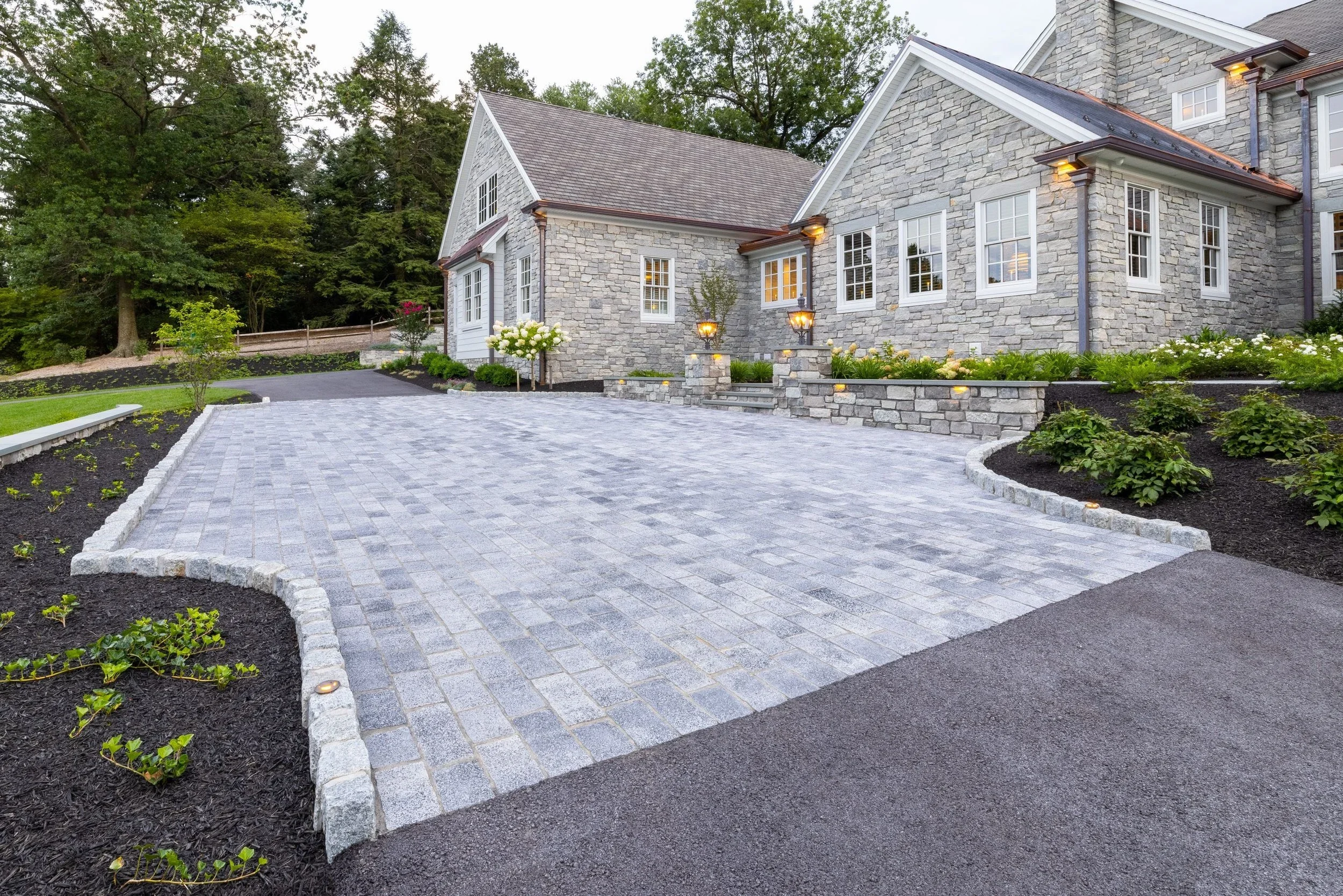 A paved driveway in front of the house is bordered by landscaped garden beds with plants and flowers.