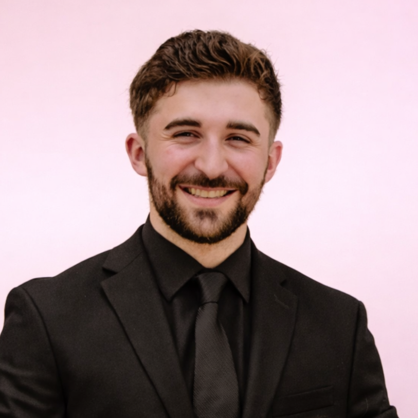 A smiling man with a beard, dressed in a black suit and tie, standing against a pink background.
