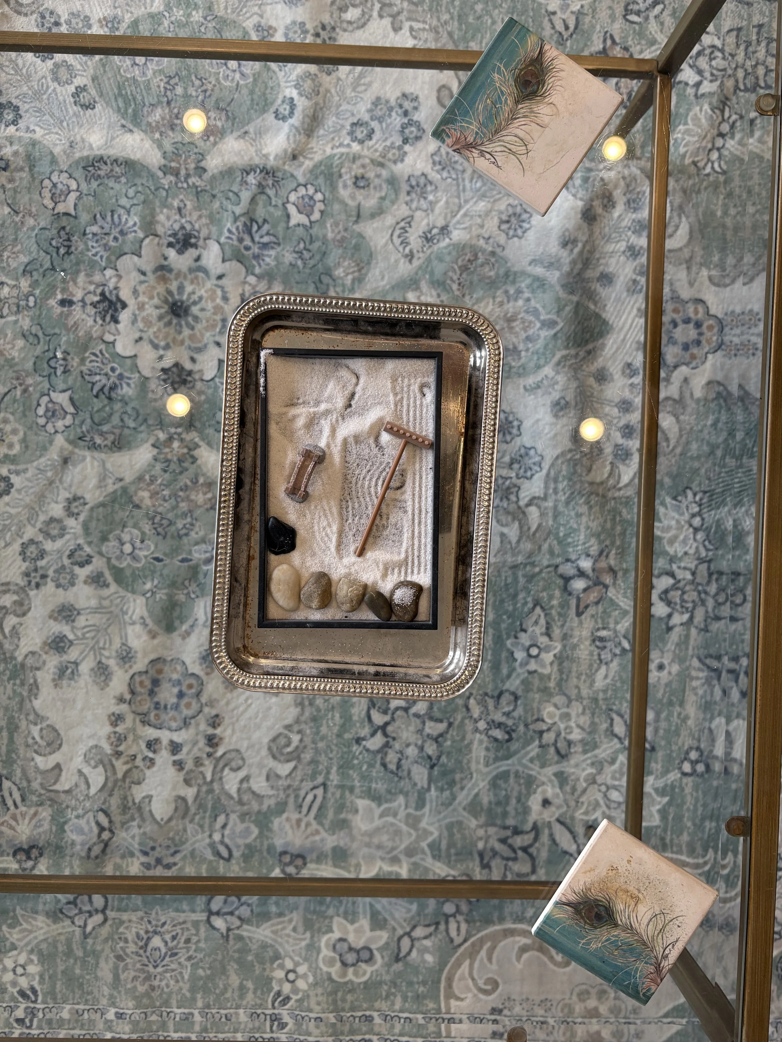 An overhead view of a glass display case with a coastal-themed sand and rock art piece featuring pebbles and sea glass, accented with two peacock feather images on small square panels at the top and bottom.