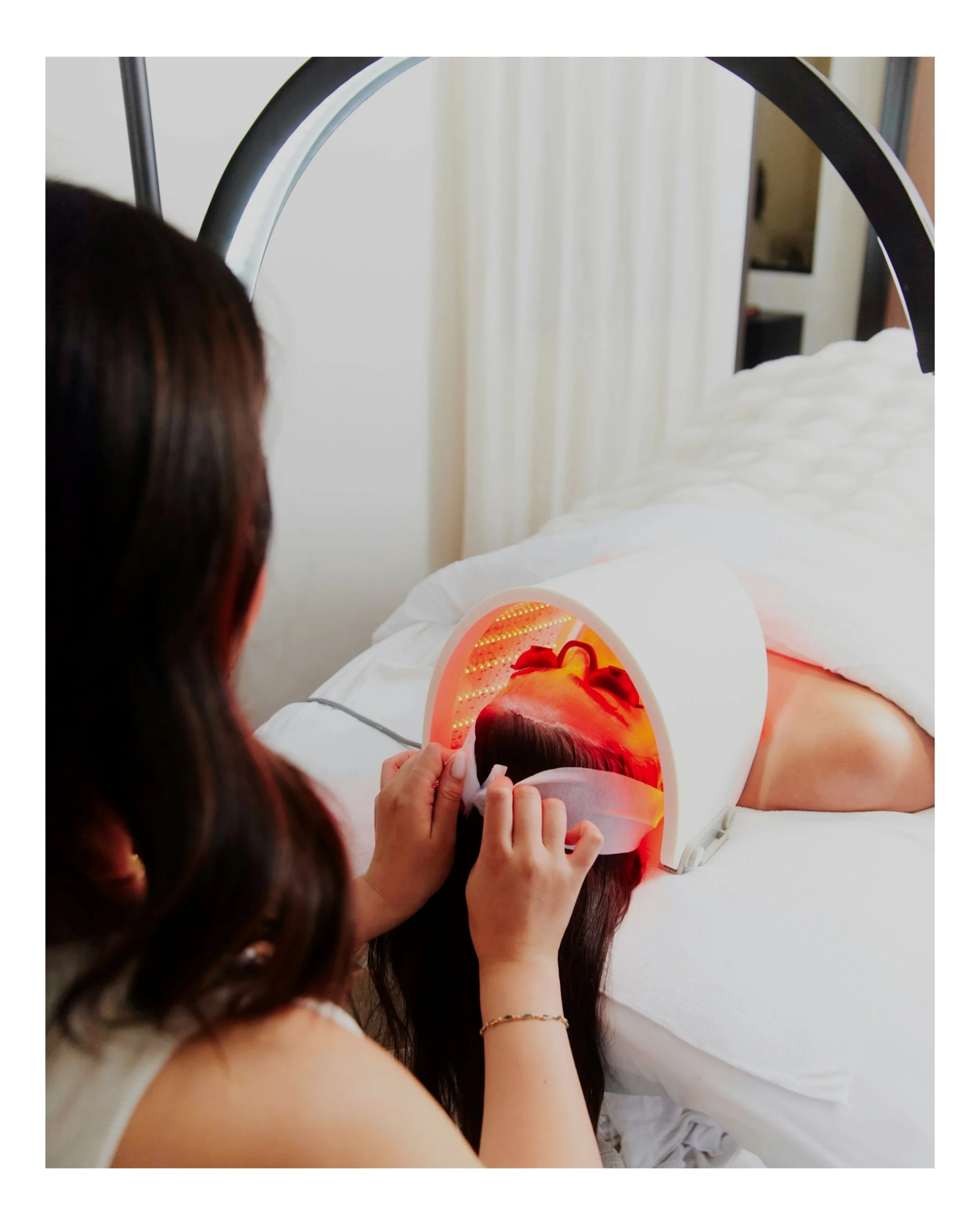 A woman undergoing a red light therapy session while lying on a bed, with her head inside a circular light device.