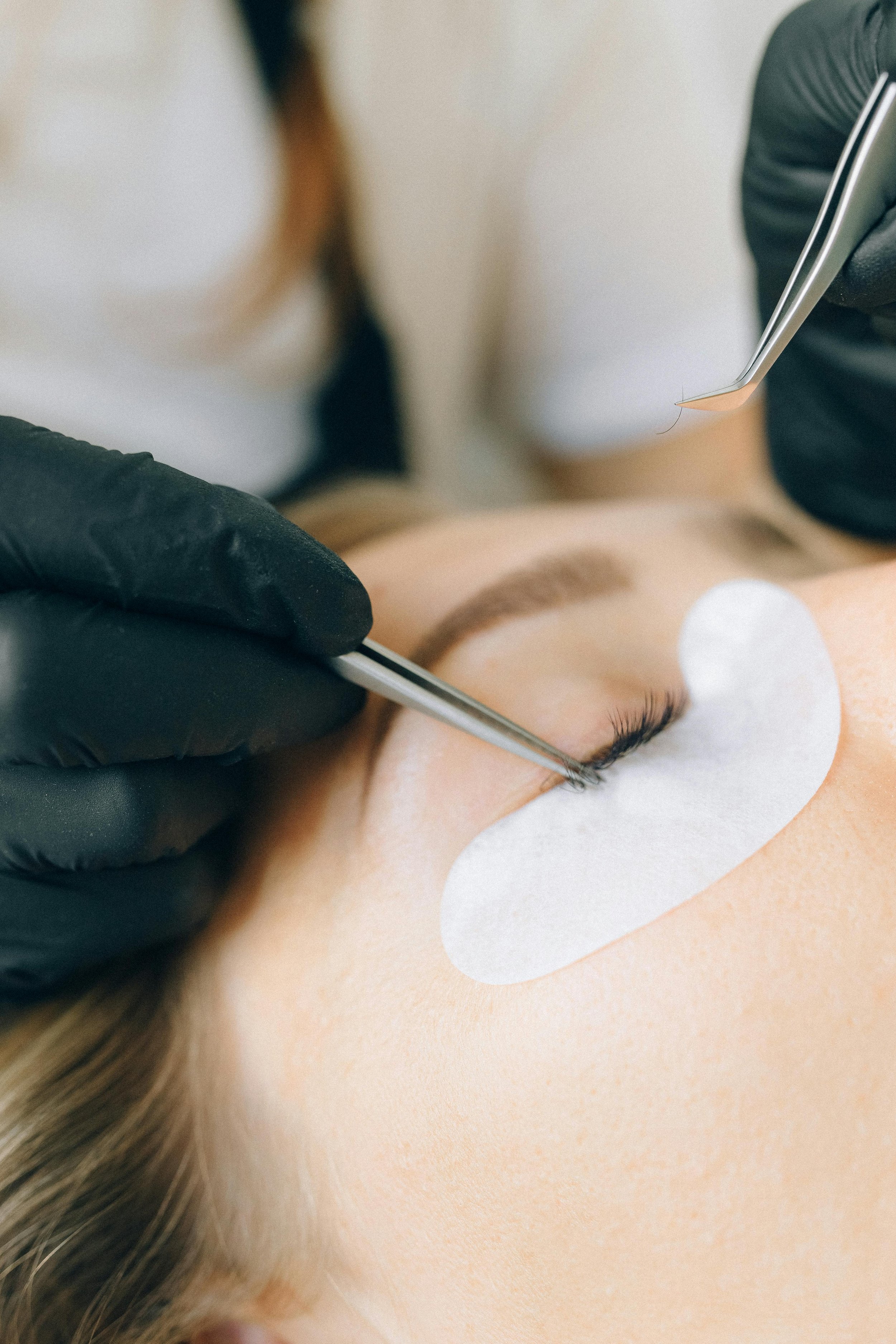 A close-up of a person receiving eyelash extension application, with a technician using tweezers and wearing black gloves, and a protective pad under the eye.