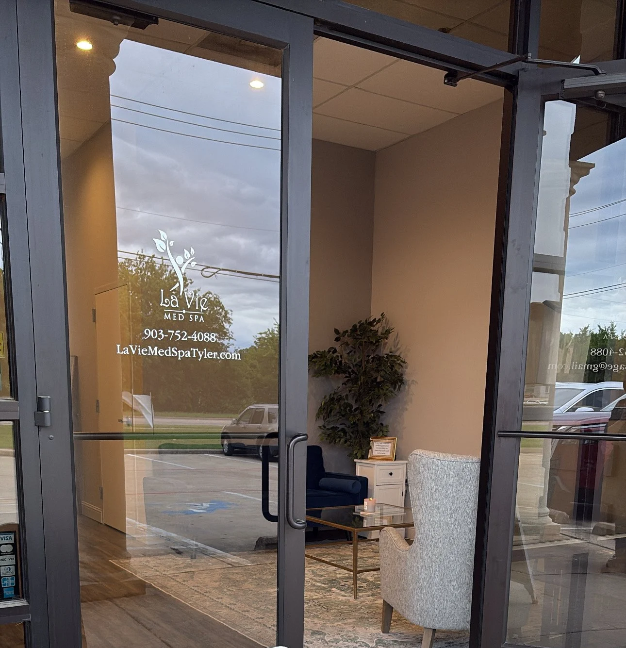 The entrance to La Vie Med Spa with glass doors, logo, and contact information handwritten on the glass, showing a waiting area inside with chairs, a table, and a plant.