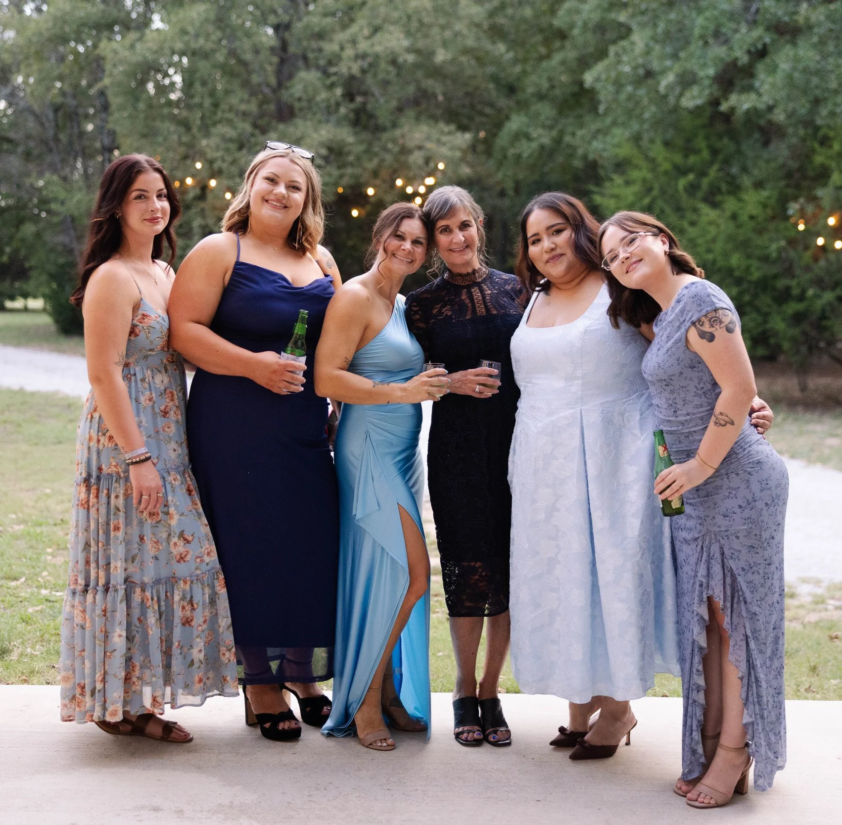 Group of six women standing outdoors dressed in formal or semi-formal attire, holding drinks, smiling for a photo.
