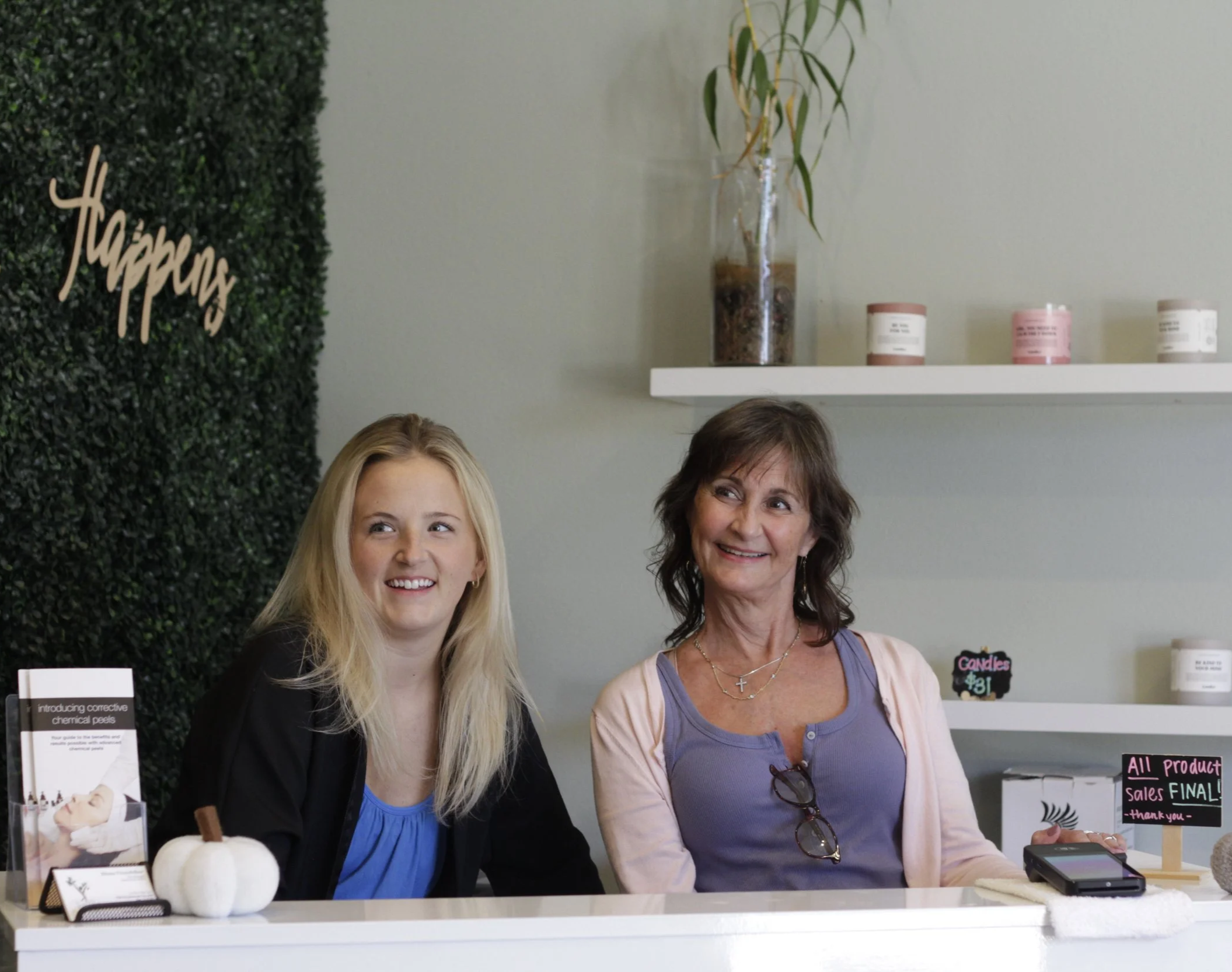 Two women sitting behind a white counter in a store, smiling. The younger woman has long blonde hair and is wearing a black blazer over a blue top. The older woman has shoulder-length brown hair and is wearing a purple top with a light pink cardigan, with glasses hanging from her neck. A small white pumpkin and a product display are on the counter, and a wall with a green leafy panel and a white shelf with candles and a tall glass vase with plant are in the background.