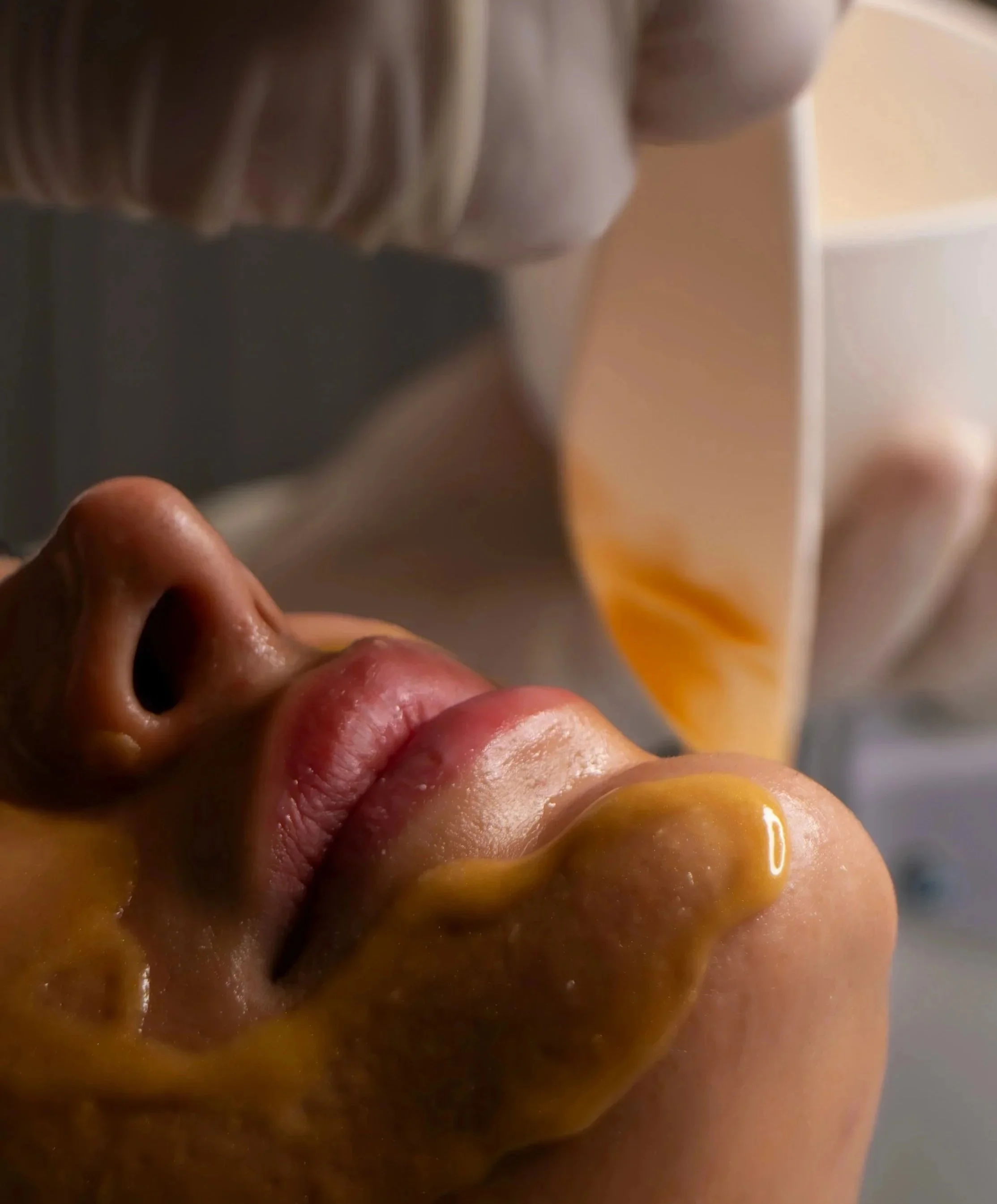 Close-up of a person lying down with their mouth slightly open, with a brownish substance near their lips and nose, and a white and orange object, possibly a cloth or blanket, covering part of their face.