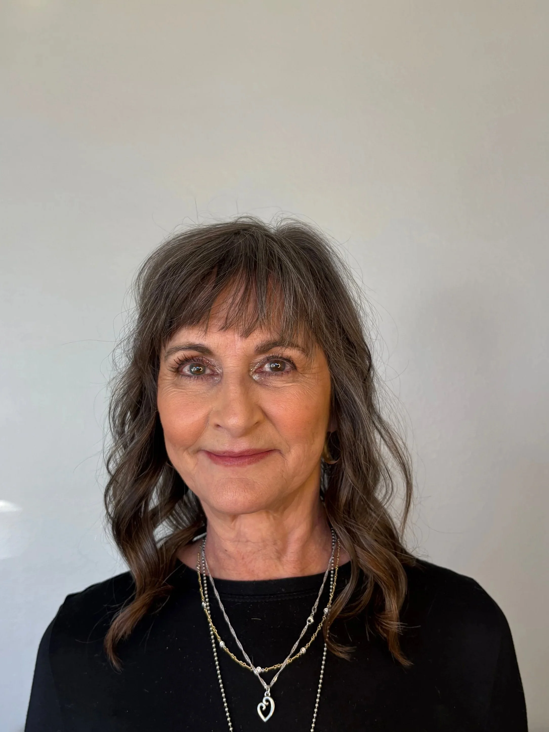 A mature woman with brown wavy hair and layered bangs, wearing gold eye makeup and a black top, accessorized with multiple layered silver necklaces, smiling against a plain light-colored background.