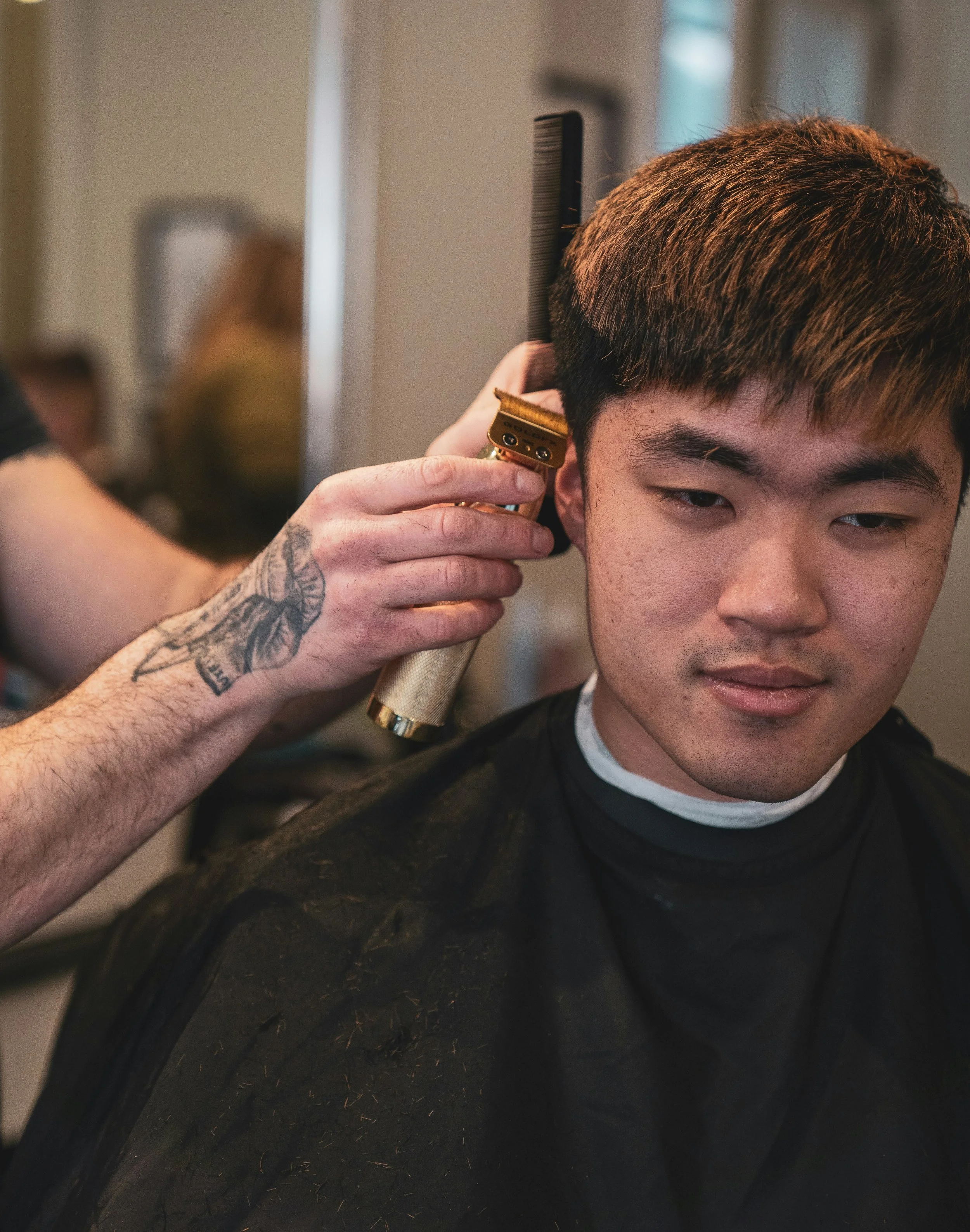 A young man getting a haircut with hair clippings on his black cape in a barbershop.