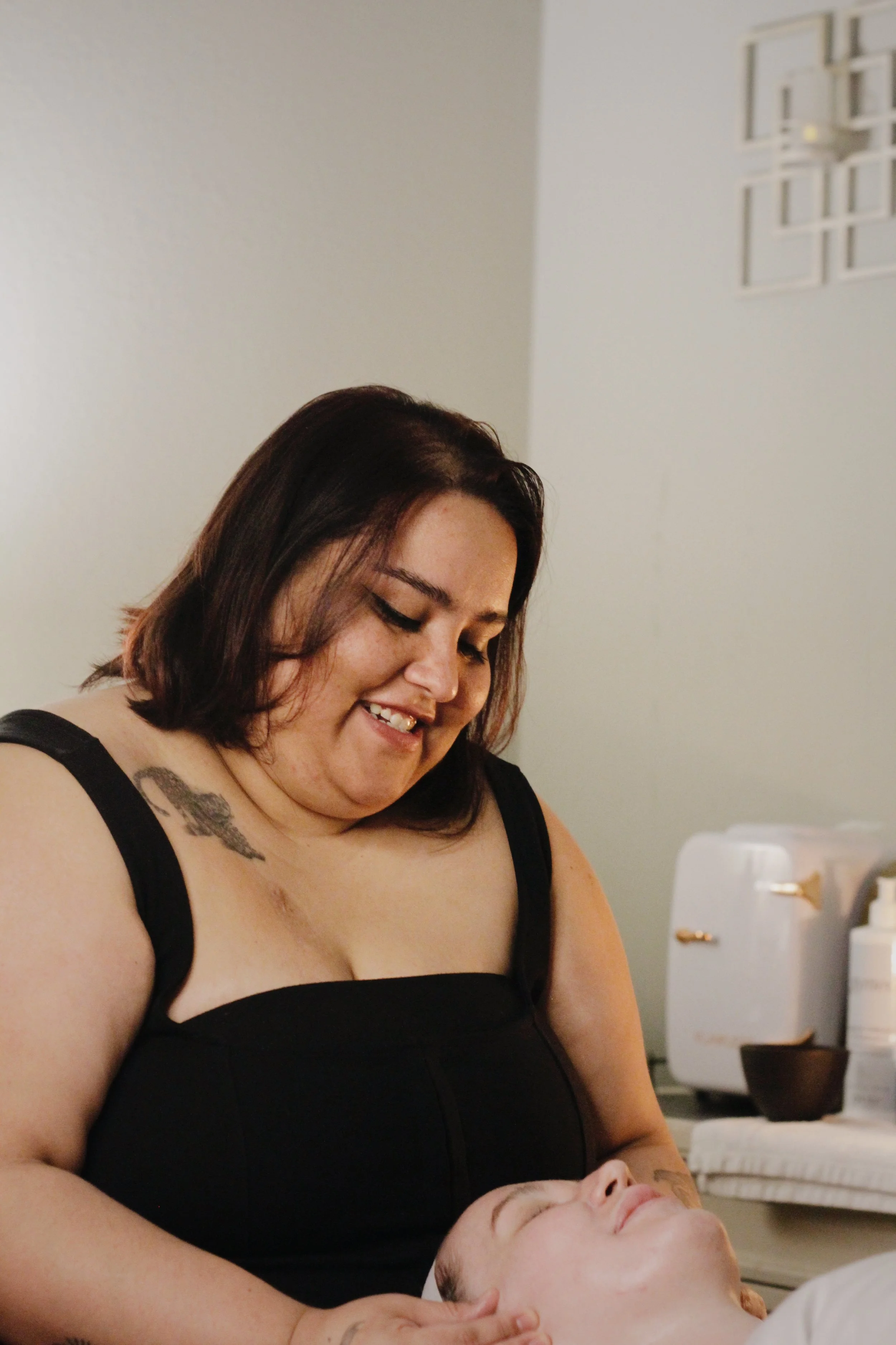 A woman with shoulder-length dark hair smiling and looking down at a person lying down with their eyes closed. The woman is wearing a black dress with thick straps and has a tattoo on her right shoulder. The background includes a white wall, some skincare or medical products, and a small black bowl.