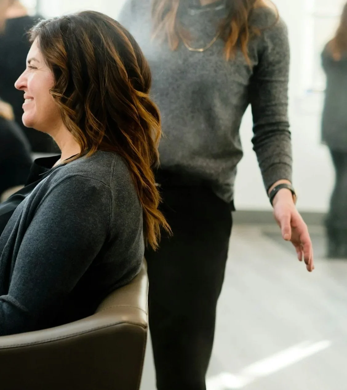 A woman with long, wavy brown hair smiling while sitting in a chair, and another person standing next to her with their hand extended slightly downward, wearing a gray sweater and black pants.