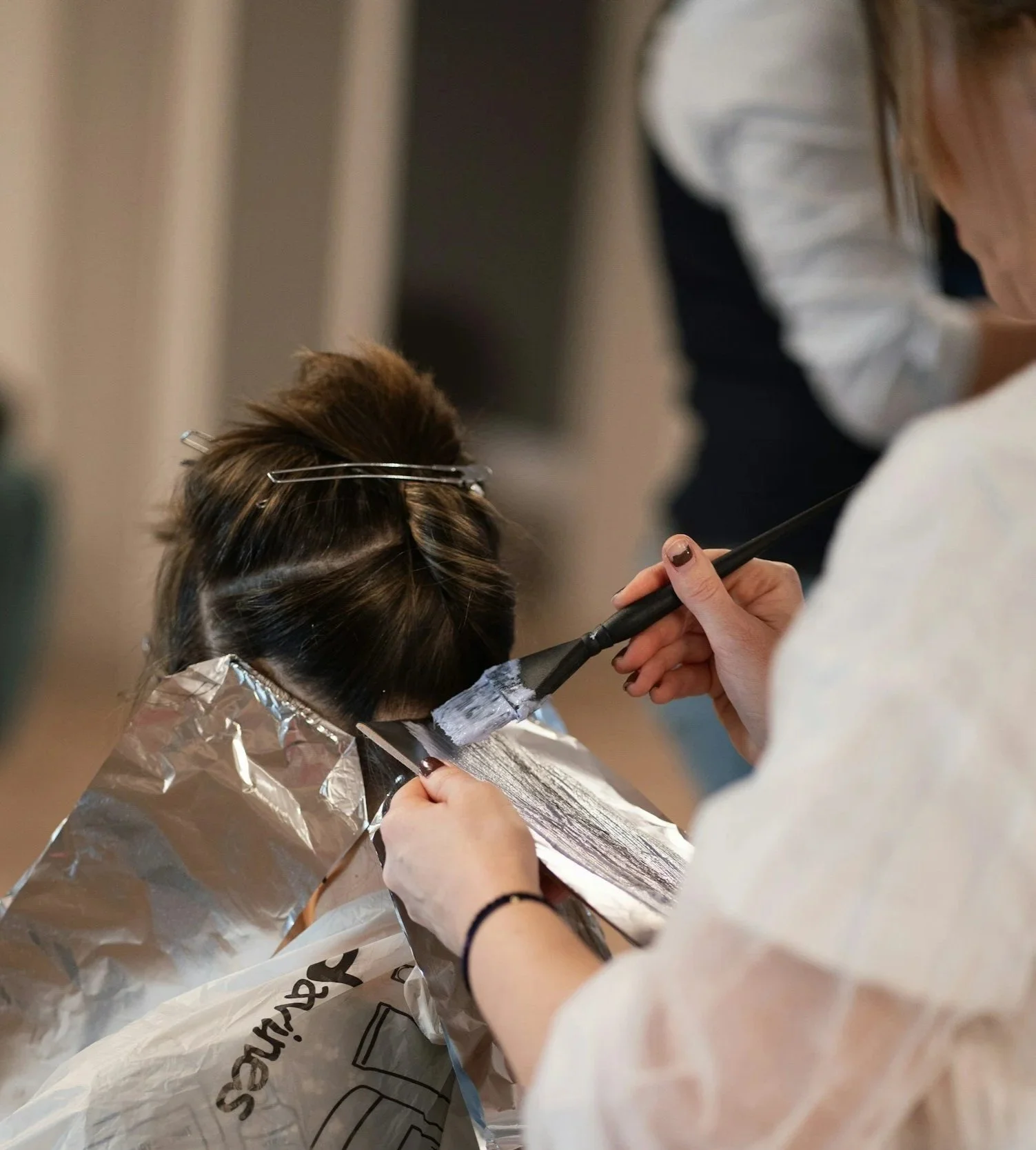 Person receiving a hair coloring treatment at a salon, with hair sectioned and foil in place, as a stylist applies color with a brush.