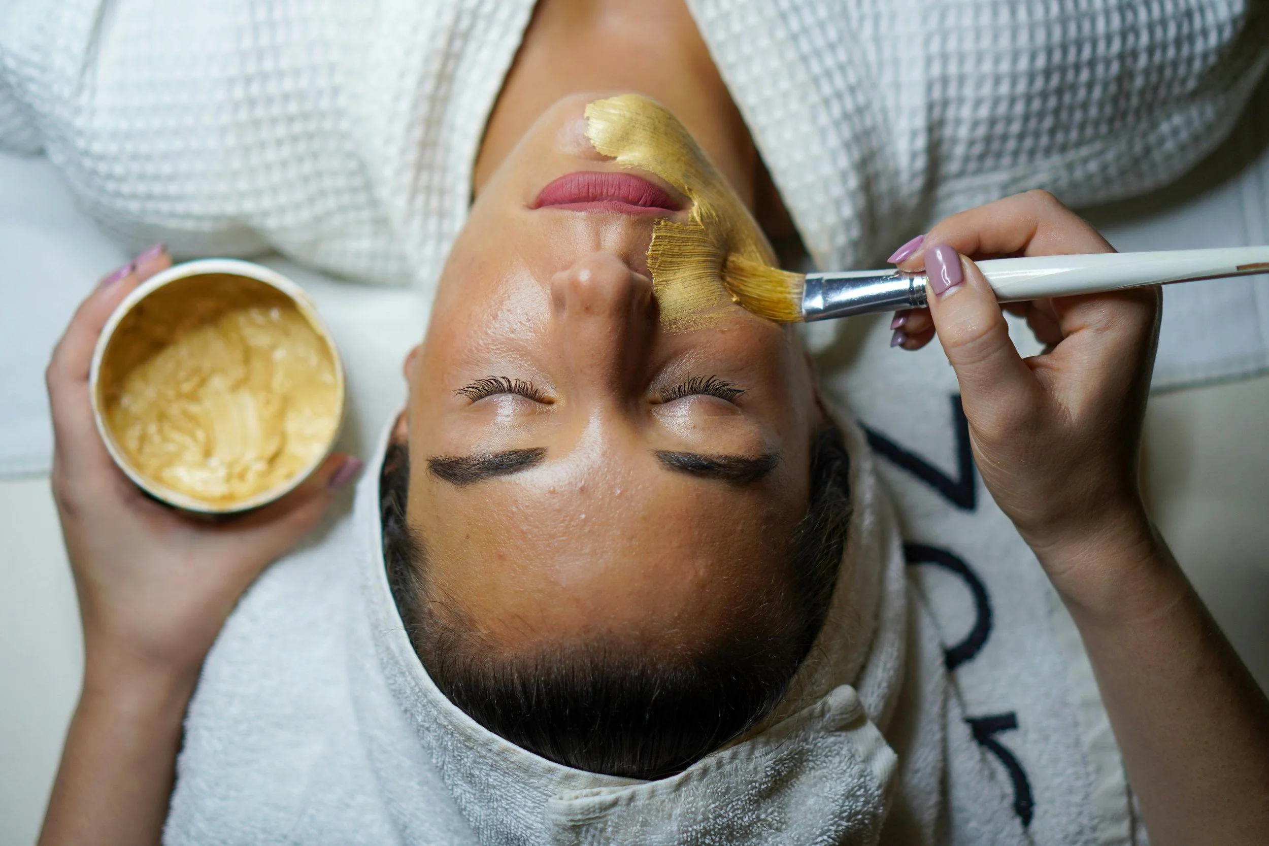 A woman lying down with her eyes closed, receiving a facial treatment. A skincare professional applies a golden facial mask with a brush, holding a bowl of the mask product.