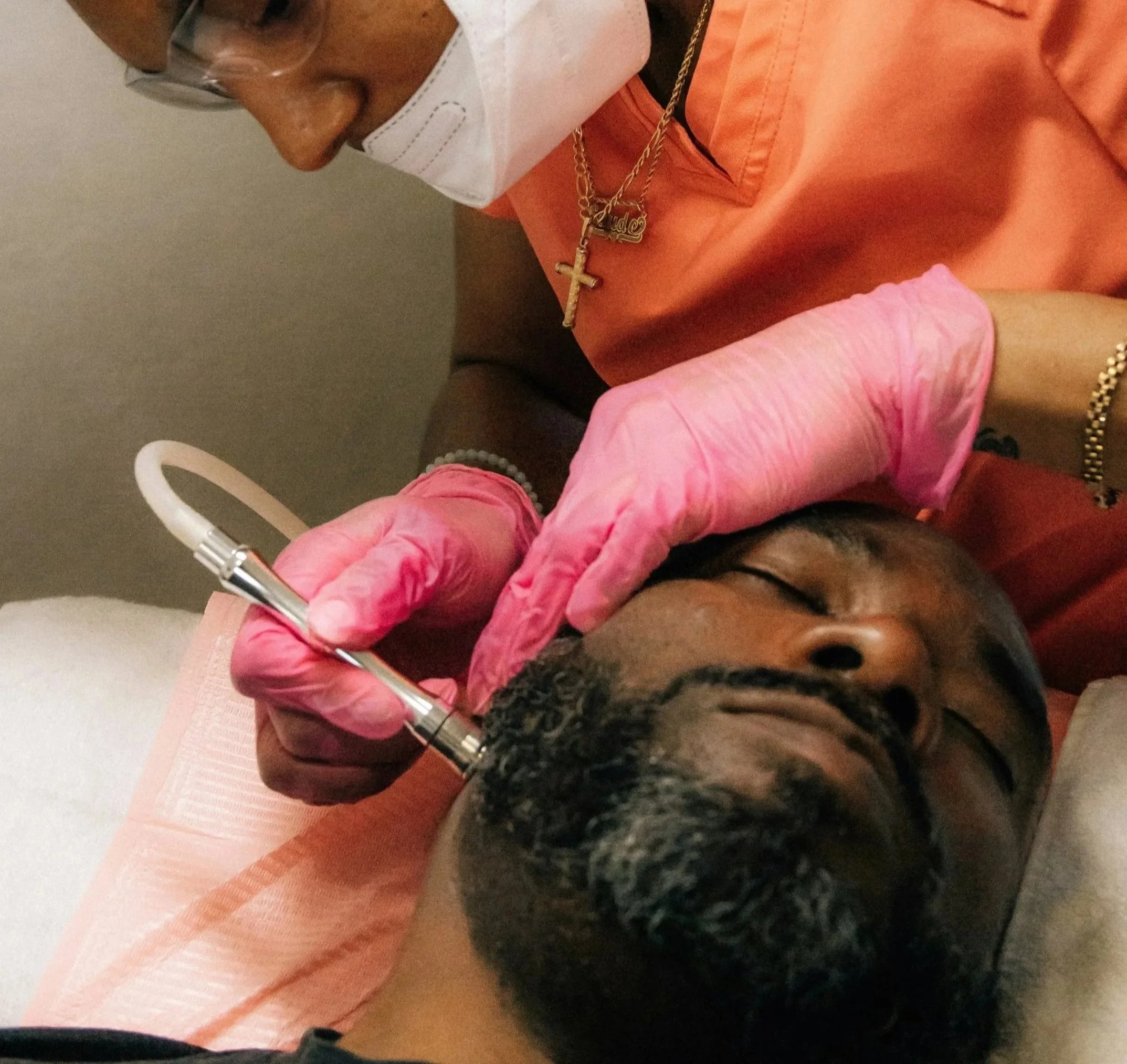 A woman in pink gloves is performing a cosmetic procedure on a man's face, who is lying down with his eyes closed. The woman is wearing a mask, glasses, and jewelry, including a gold chain with a cross pendant.