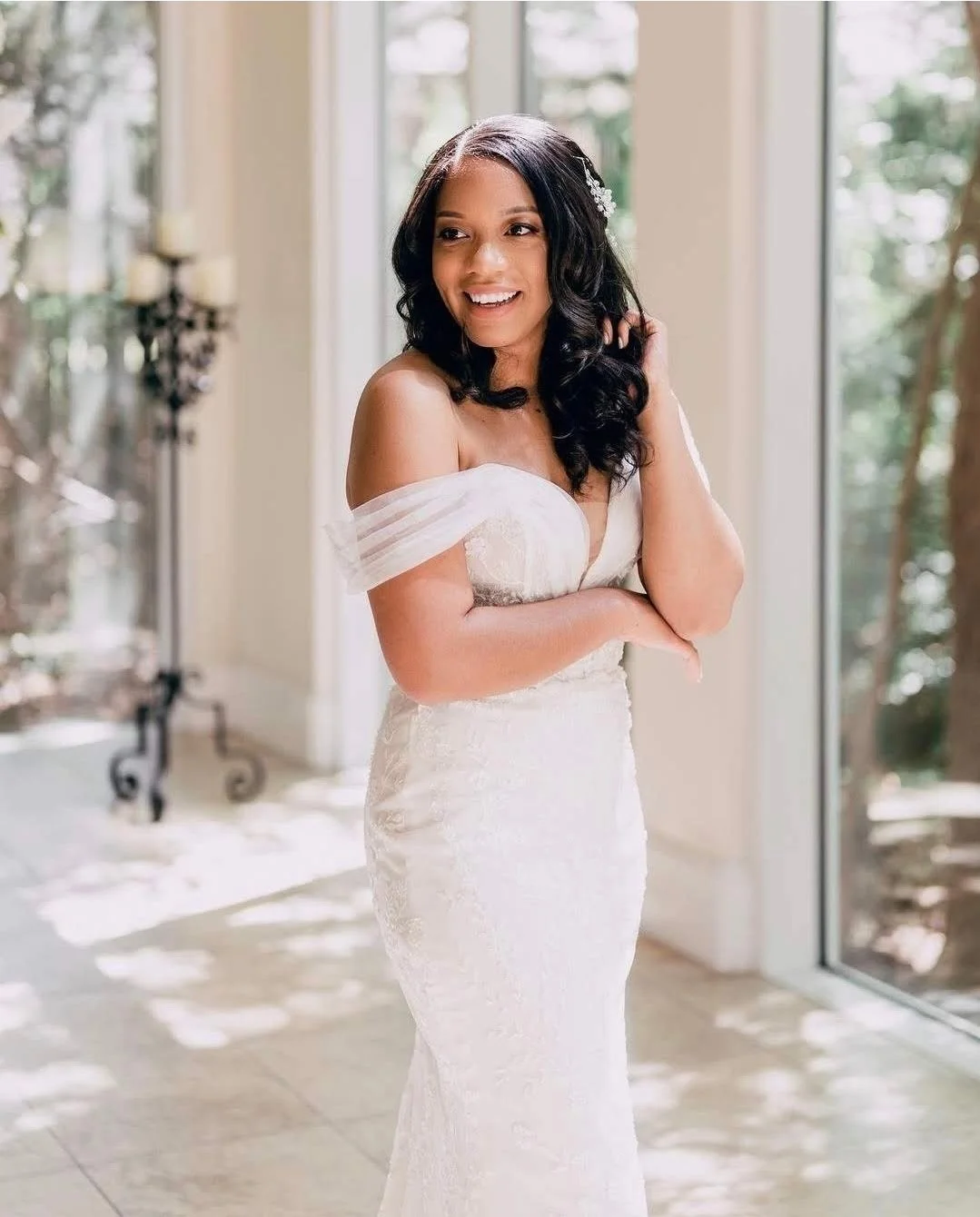A woman in a wedding dress standing in front of large windows with sunlight streaming in, smiling and touching her hair.