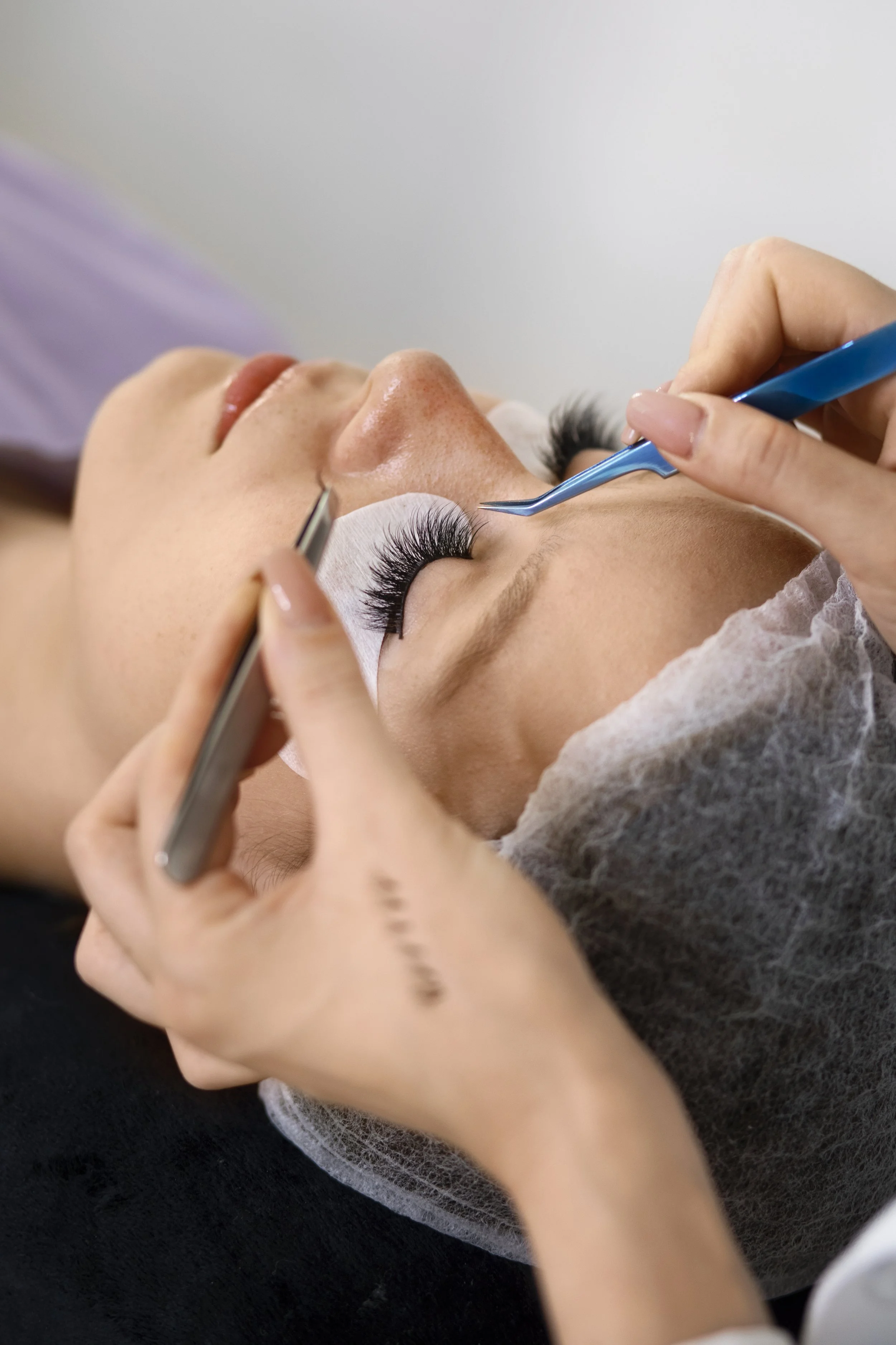 A person lying down with eyes closed, receiving eyelash extensions from a beauty technician using tweezers, with the technician working with a small tool in their right hand.