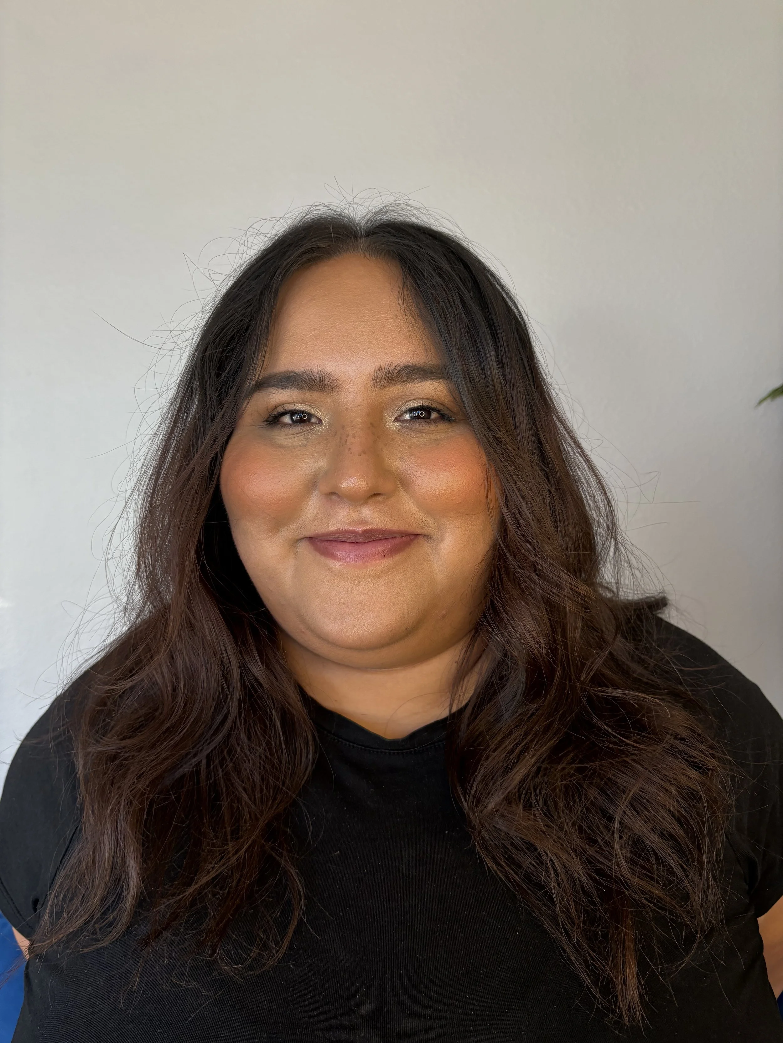 A woman with long dark hair and light makeup, wearing a black top, smiling against a plain light gray background.