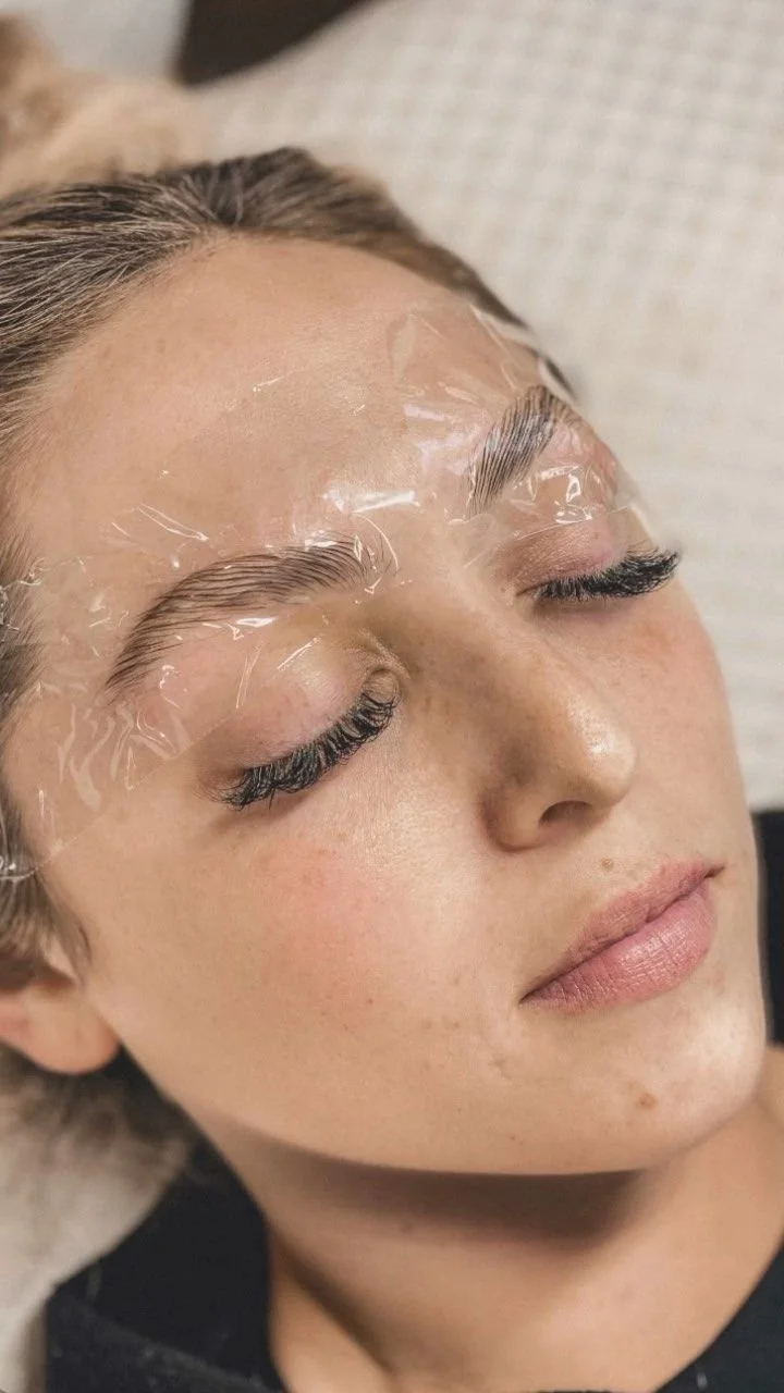 A woman with closed eyes receiving a cosmetic treatment, with her eyebrows covered in clear plastic wrap.