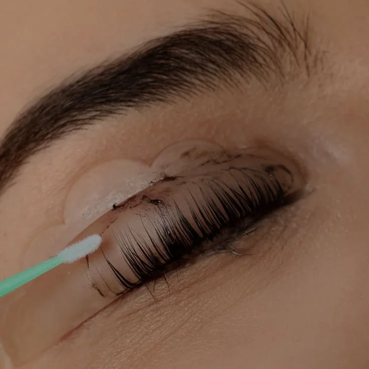 Close-up of a person's closed eye with long eyelashes, showing an eyelash extension application process with a cotton swab near the eye.