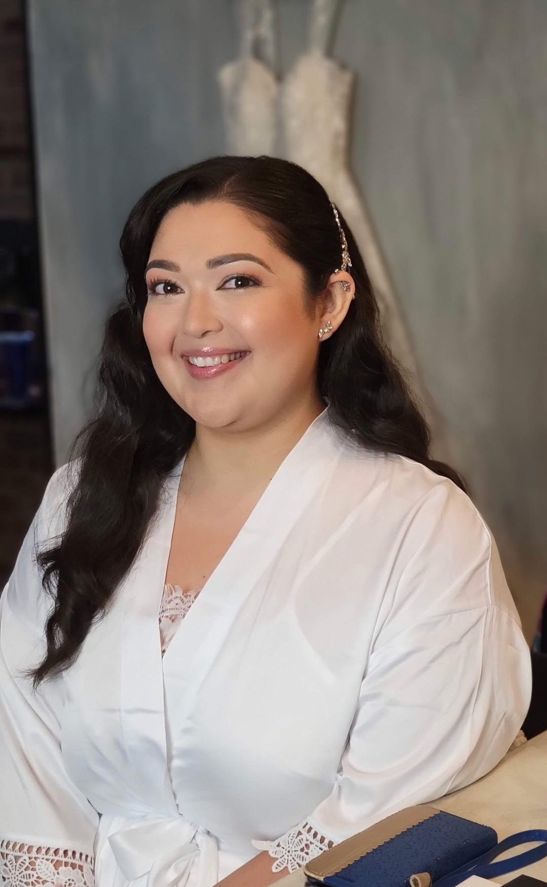 A woman with dark wavy hair dressed in a white robe, smiling, in front of a blurred background with a dress form.