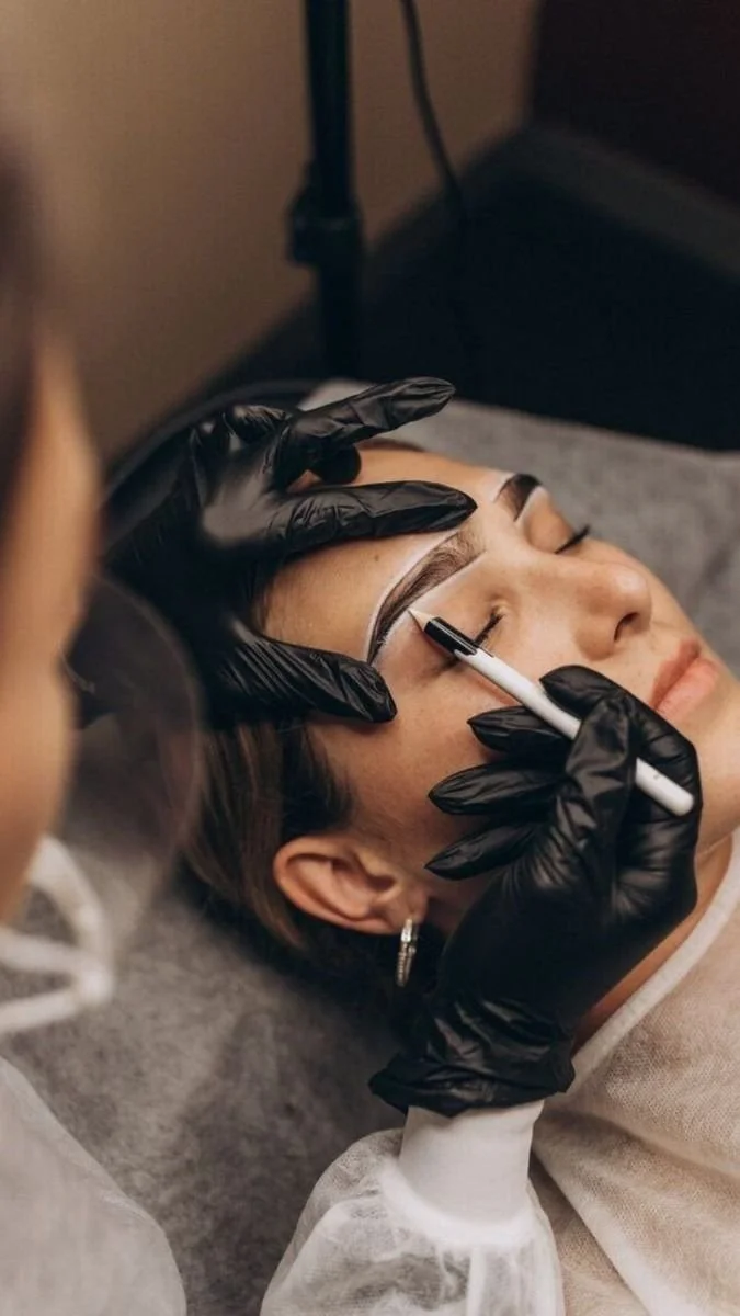 A woman lying down while a technician applies permanent makeup to her eyebrows using a pen-like device. The technician is wearing black gloves.