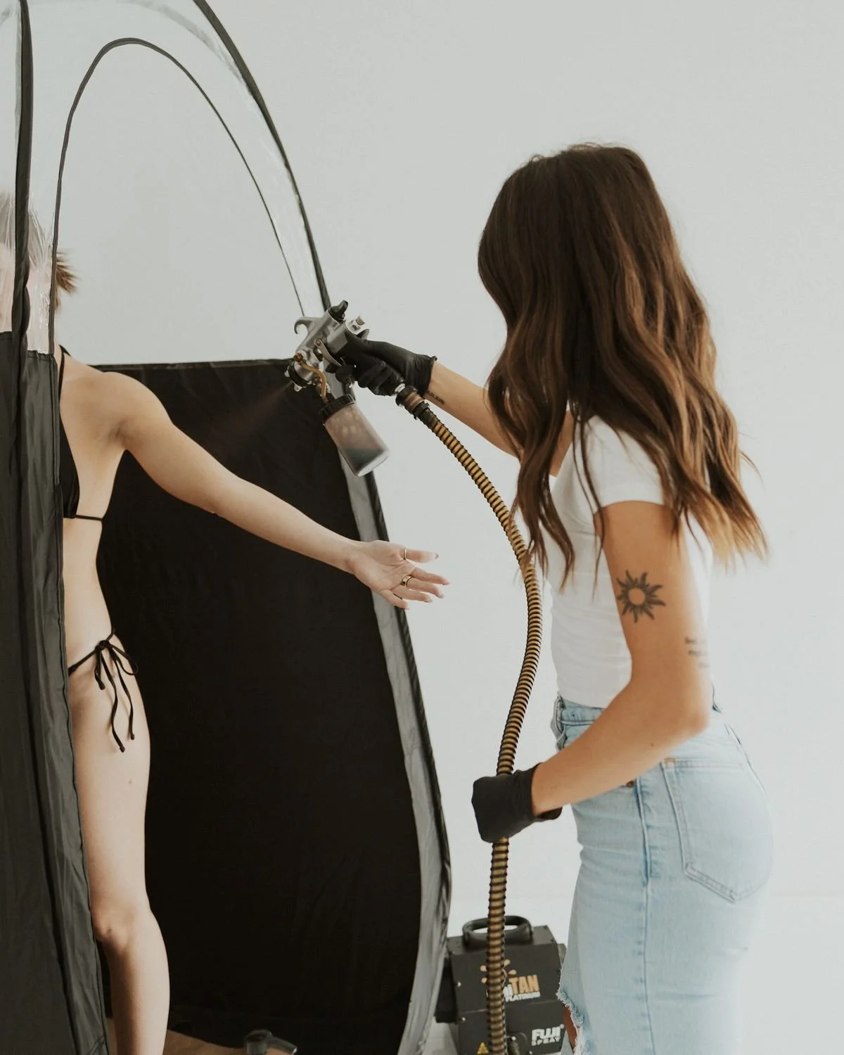 Person using spray gun to apply paint or makeup to another person’s exposed arm, with black tent-like privacy divider and equipment in the background.