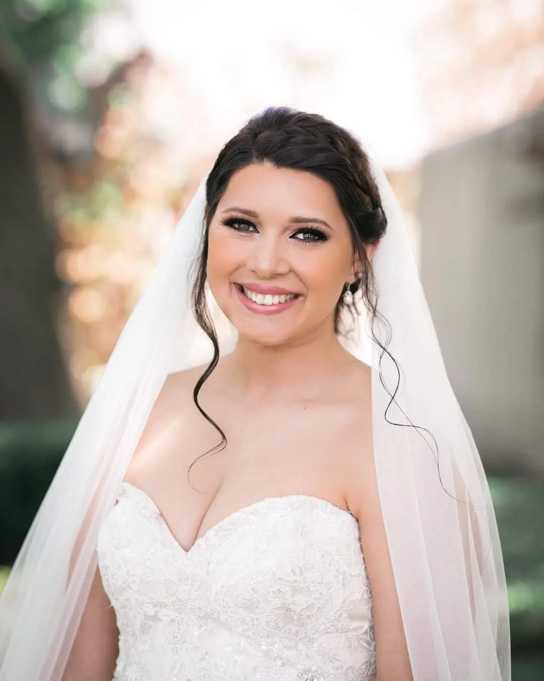 A bride smiling outdoors in a white wedding dress and veil with dark hair and makeup.