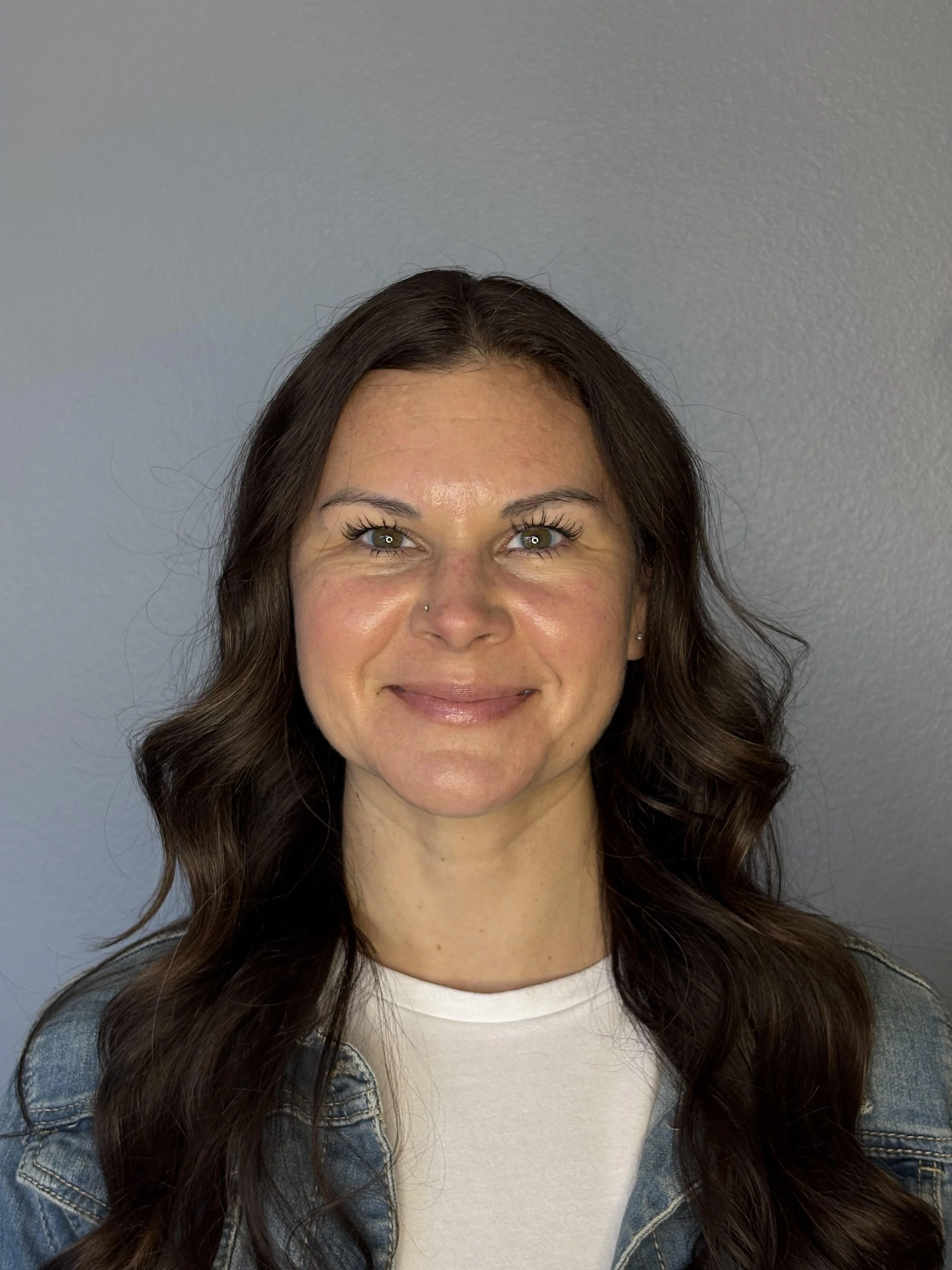 A woman with long wavy brown hair wearing a denim jacket and white shirt, smiling at the camera, standing against a plain gray wall.