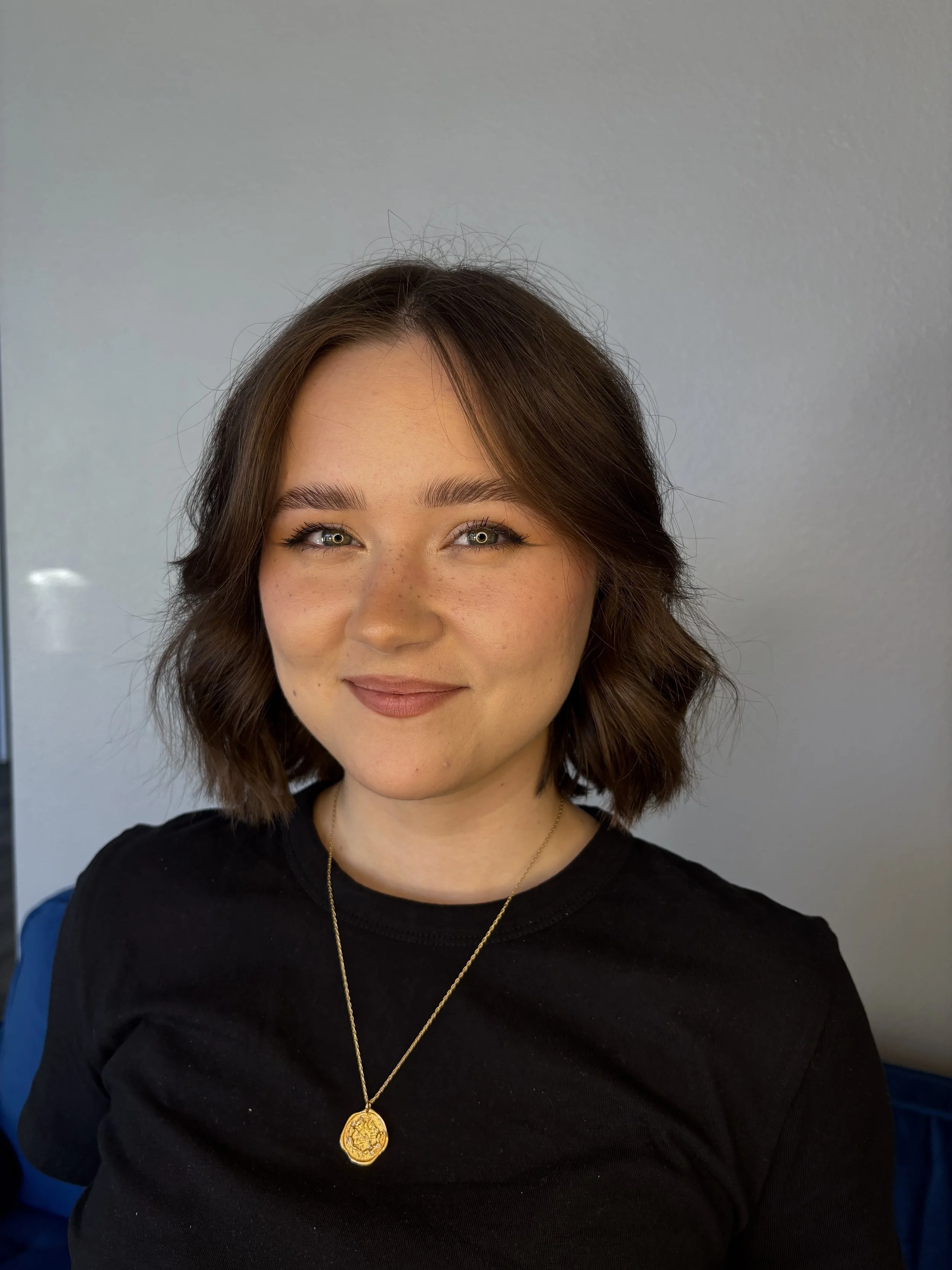 A young woman with shoulder-length brown hair, light skin, and hazel eyes, smiling, wearing a black shirt and a gold necklace with a pendant, posing against a plain gray wall.