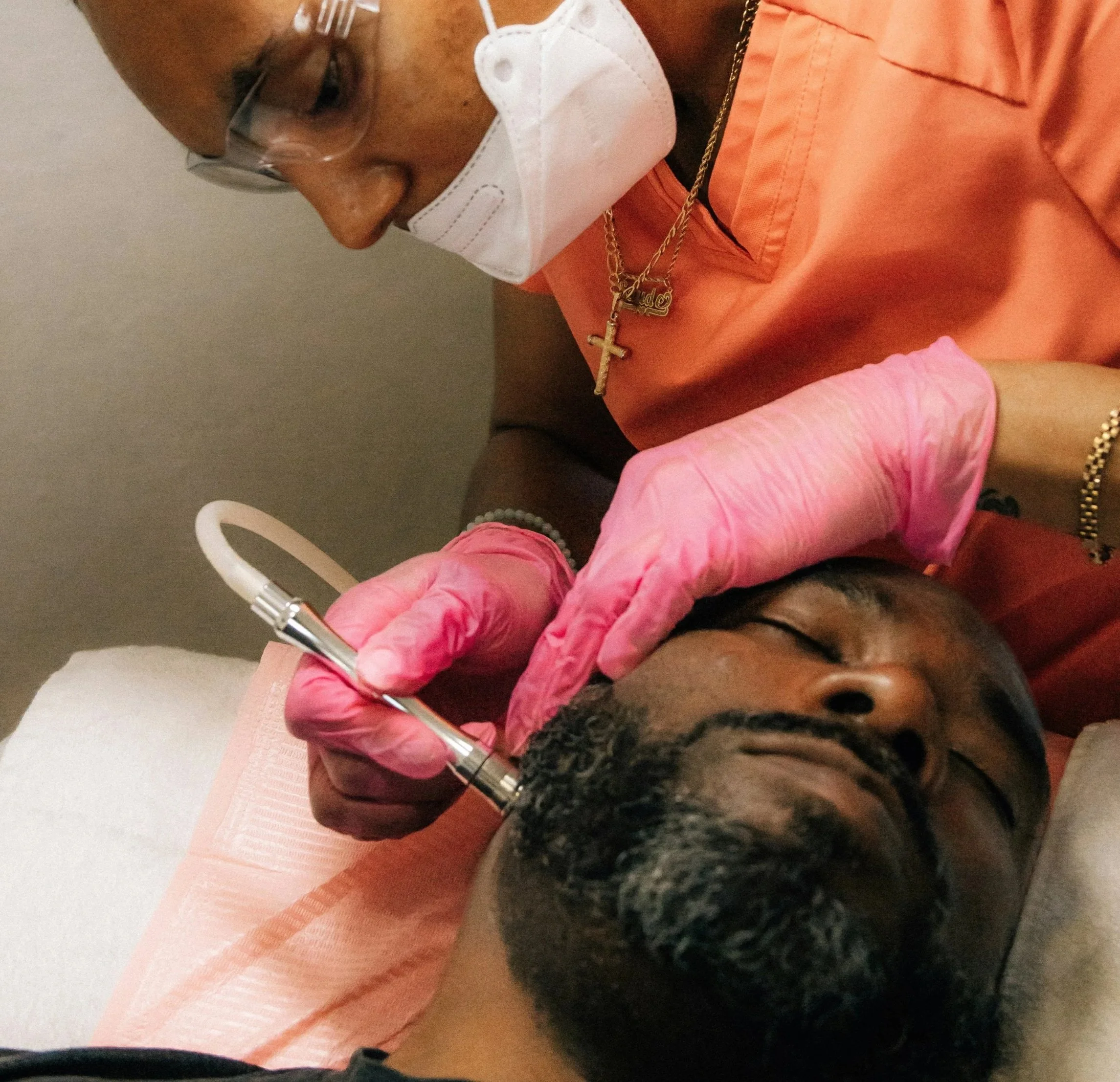 A person receiving a facial treatment from a skincare professional in a clinical setting. The professional is wearing pink gloves and a white face mask, holding a skincare instrument near the patient's face.