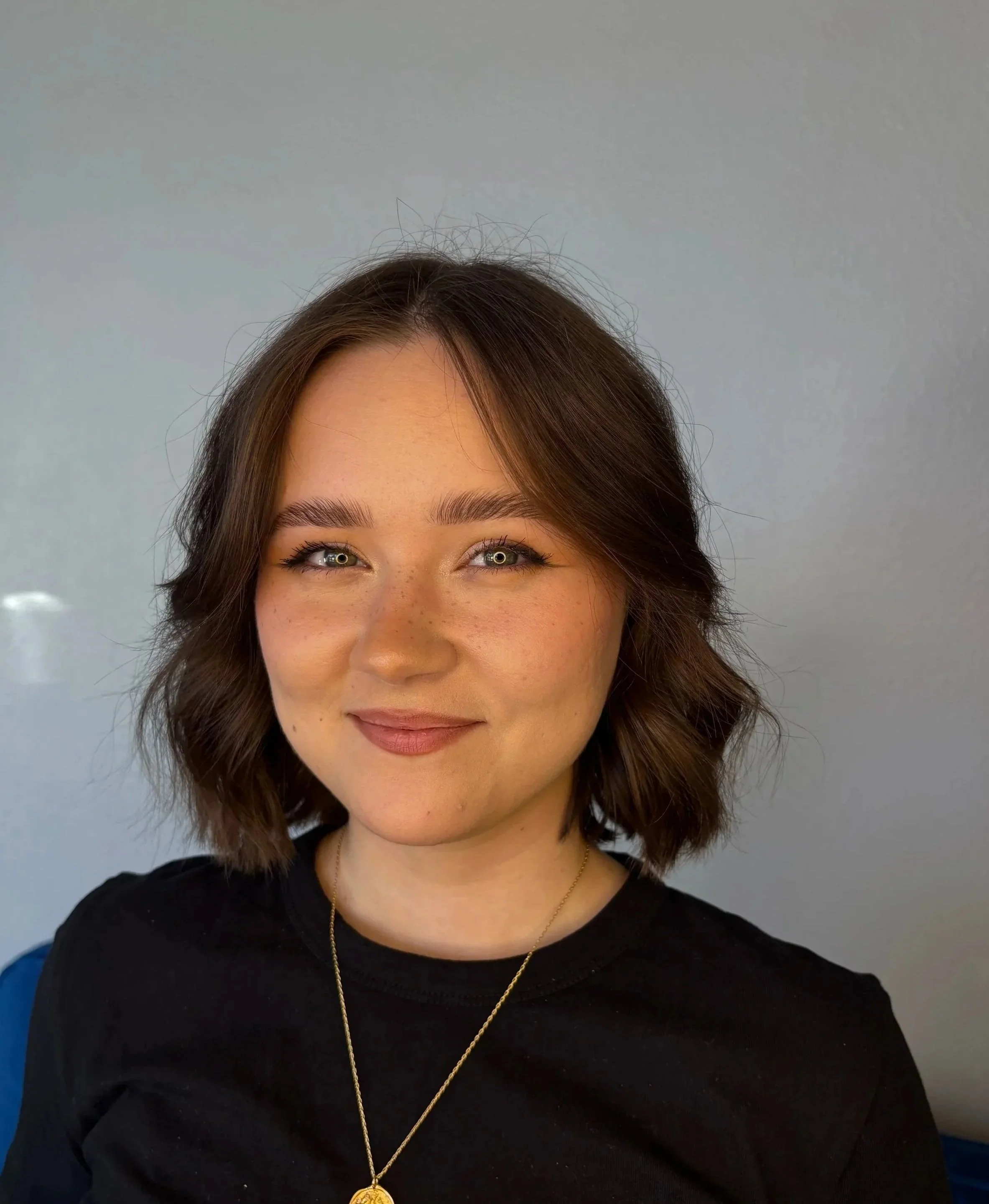 Portrait of a young woman with shoulder-length wavy brown hair, wearing a black top and a gold necklace, smiling gently, against a plain gray background.