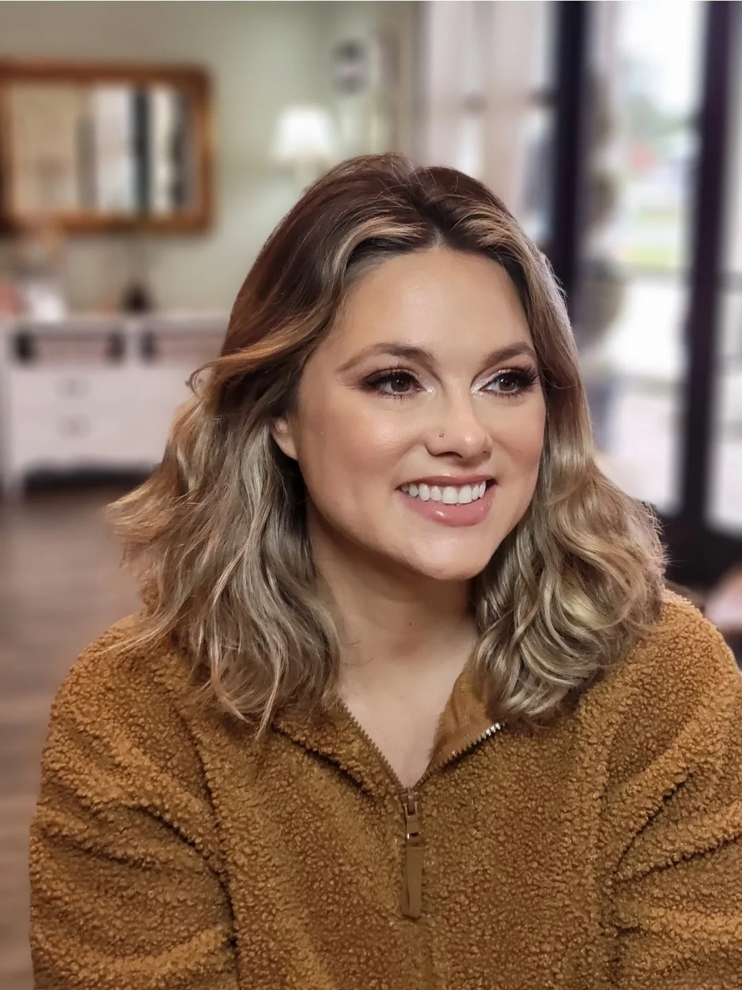 A smiling young woman with wavy brown hair and a nose piercing, wearing a brown zip-up fleece jacket, sitting inside a cozy cafe or restaurant with blurred background including a mirror, lamp, and large window.