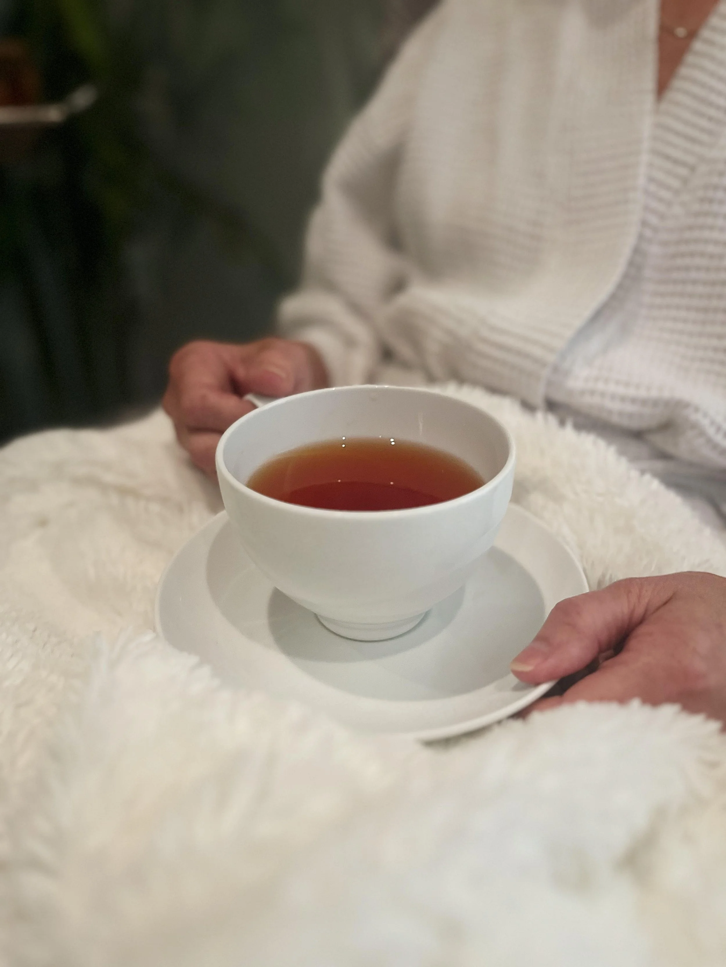 A person holding a white teacup filled with tea on a matching saucer, resting on a soft, white fuzzy surface.