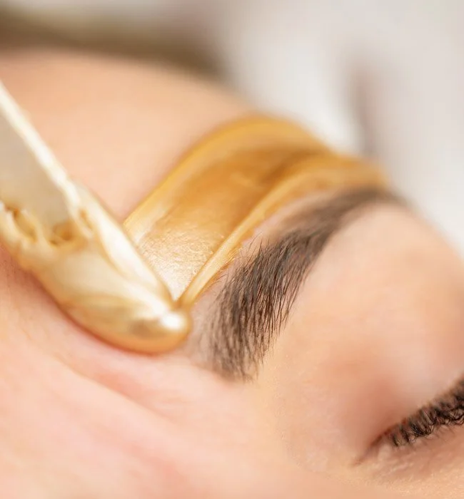 Close-up of a person receiving a brow waxing treatment, with wax being applied to the eyebrow area.