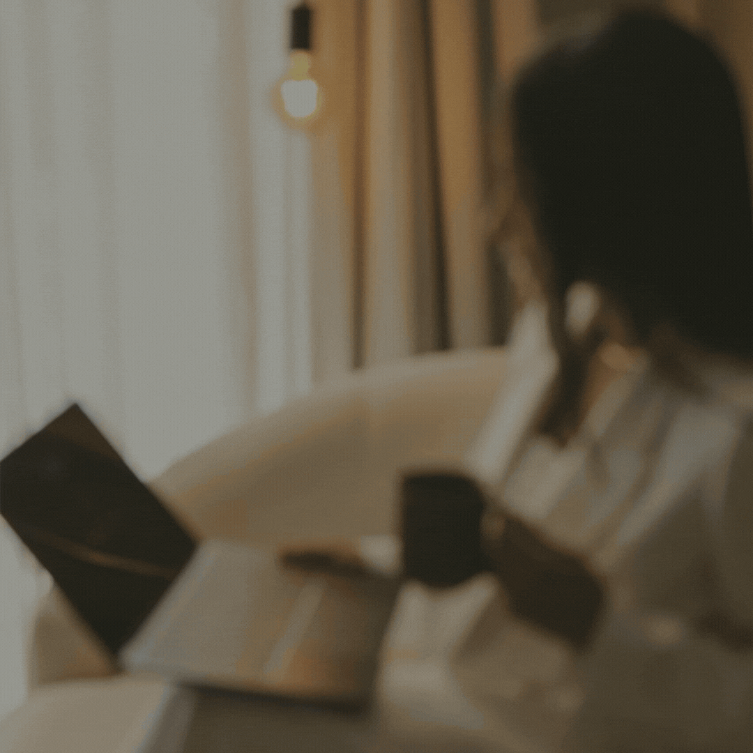 A woman sitting on a beige sofa, reading a book, with a phone, and a laptop nearby. The room is softly lit with a hanging light bulb and a mirror on the wall.