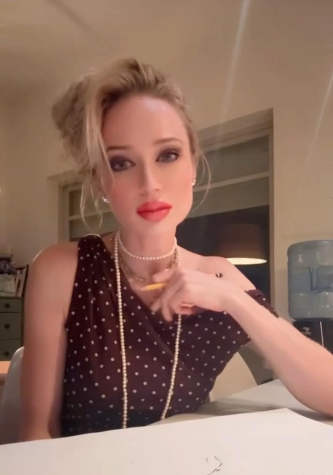 A woman with blonde hair styled up, wearing a dark brown polka dot dress, pearl jewelry, and red lipstick, sitting at a table in an indoor setting with beige walls and a window in the background.