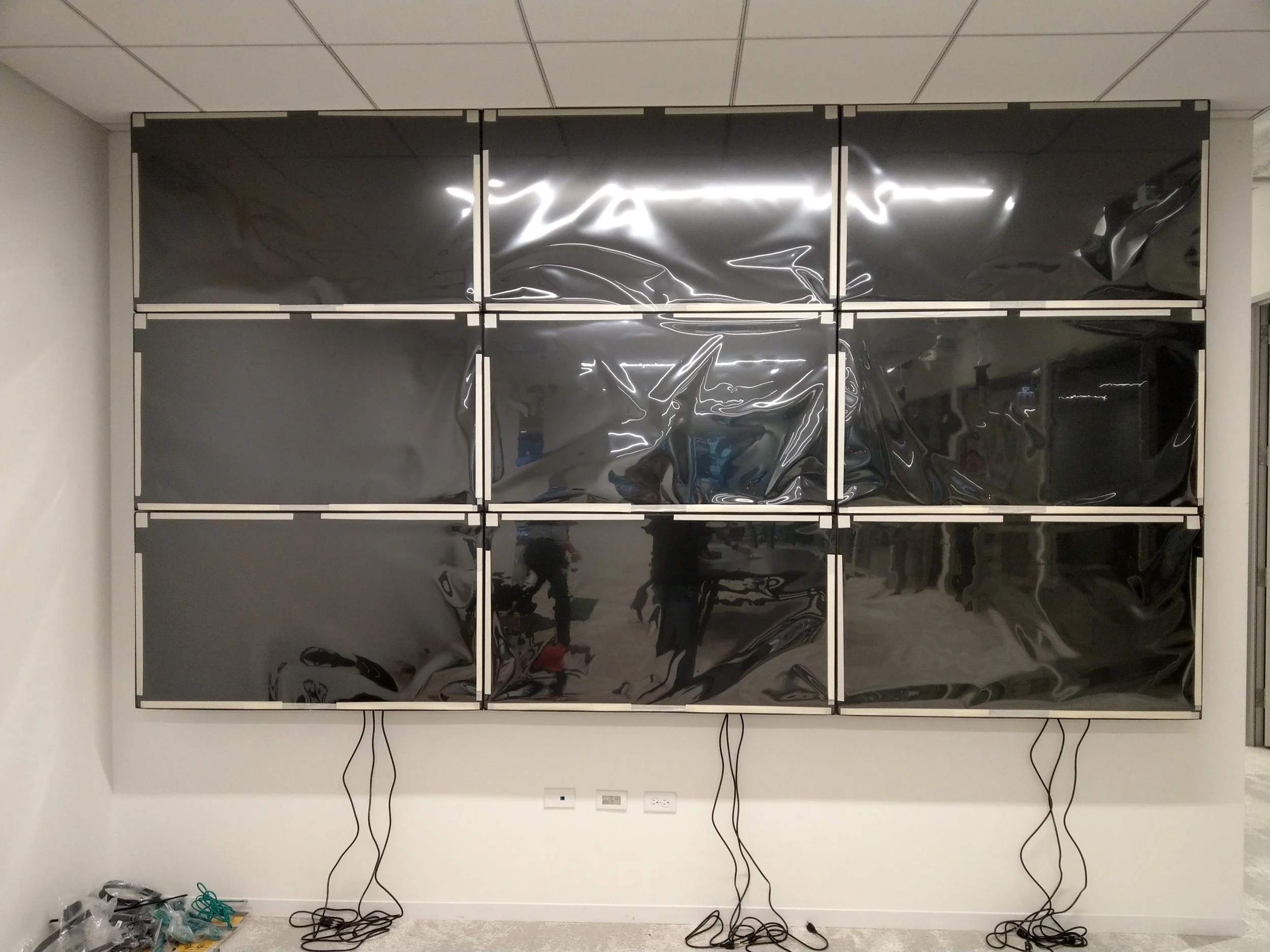 Multiple black, reflective display screens arranged in a grid mounted on a wall in an indoor space. Wires hang down from the bottom of the screens, and there are some tools or materials on the floor below.