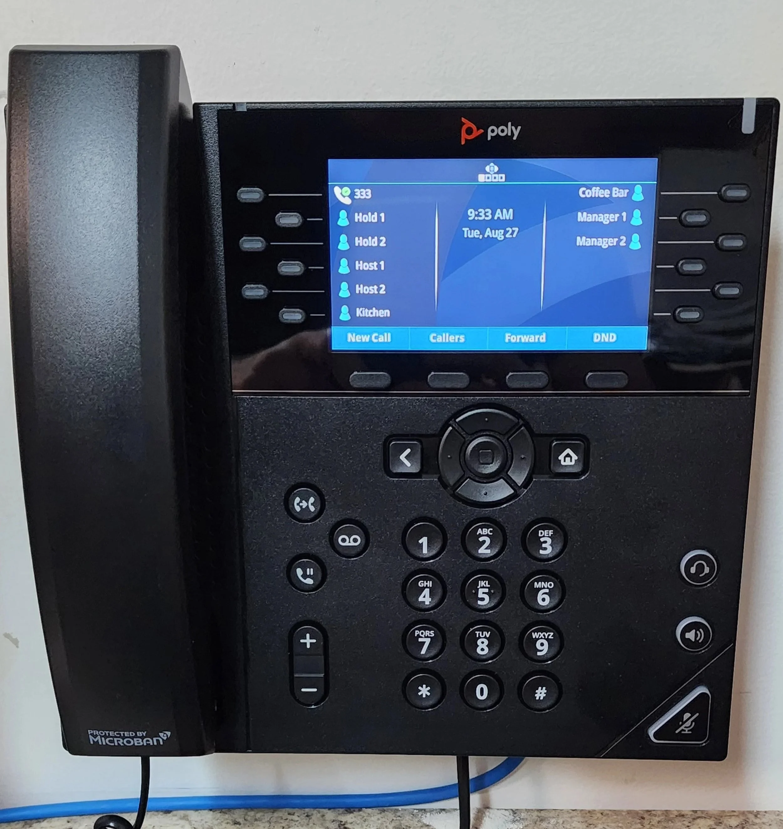 A black Poly office telephone with a screen displaying a call list, time, date, and options, mounted on a wall above a desk.