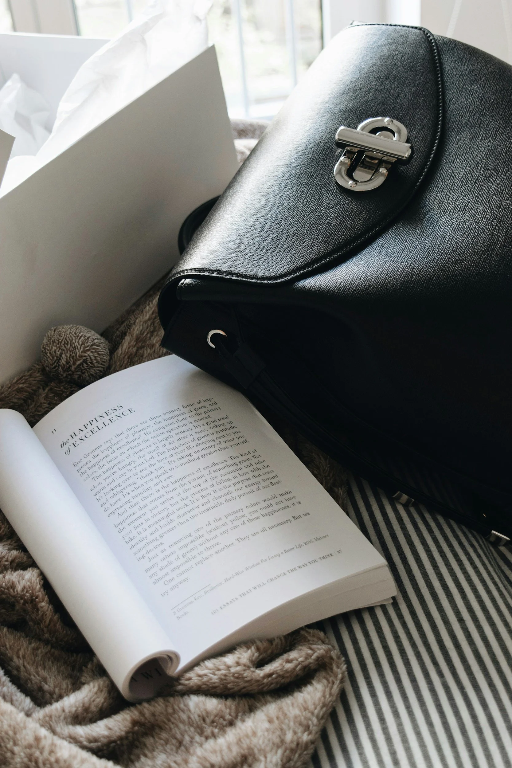 Open book with text about happiness, black leather handbag, white paper, beige blanket, and striped cloth on a bed near a window.