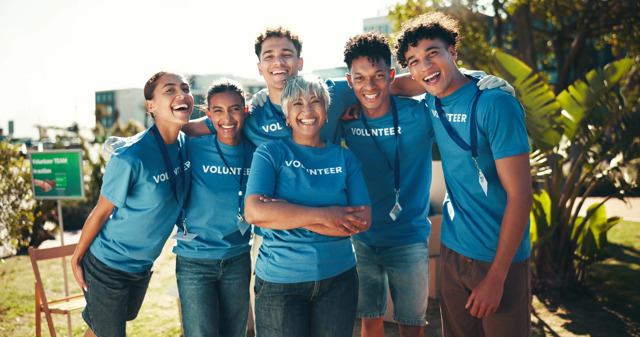 Group of six volunteers, wearing blue shirts that say 'Volunteer,' smiling and standing together outdoors on a sunny day.