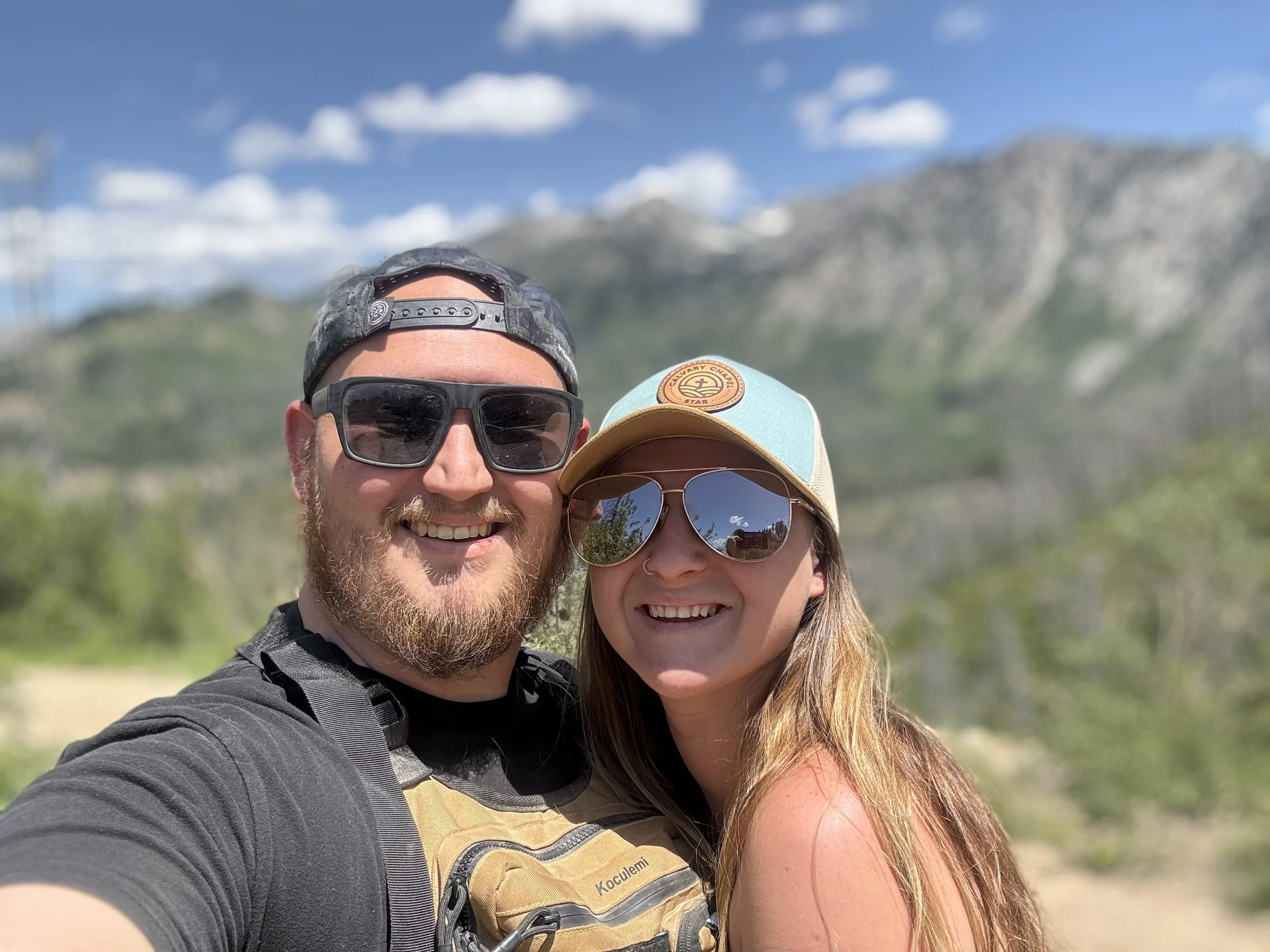 A smiling man and woman taking a selfie outdoors with mountain scenery in the background. The man has a beard, wears sunglasses, a backwards cap, and a gray shirt. The woman wears sunglasses, a baseball cap, and has long reddish hair.