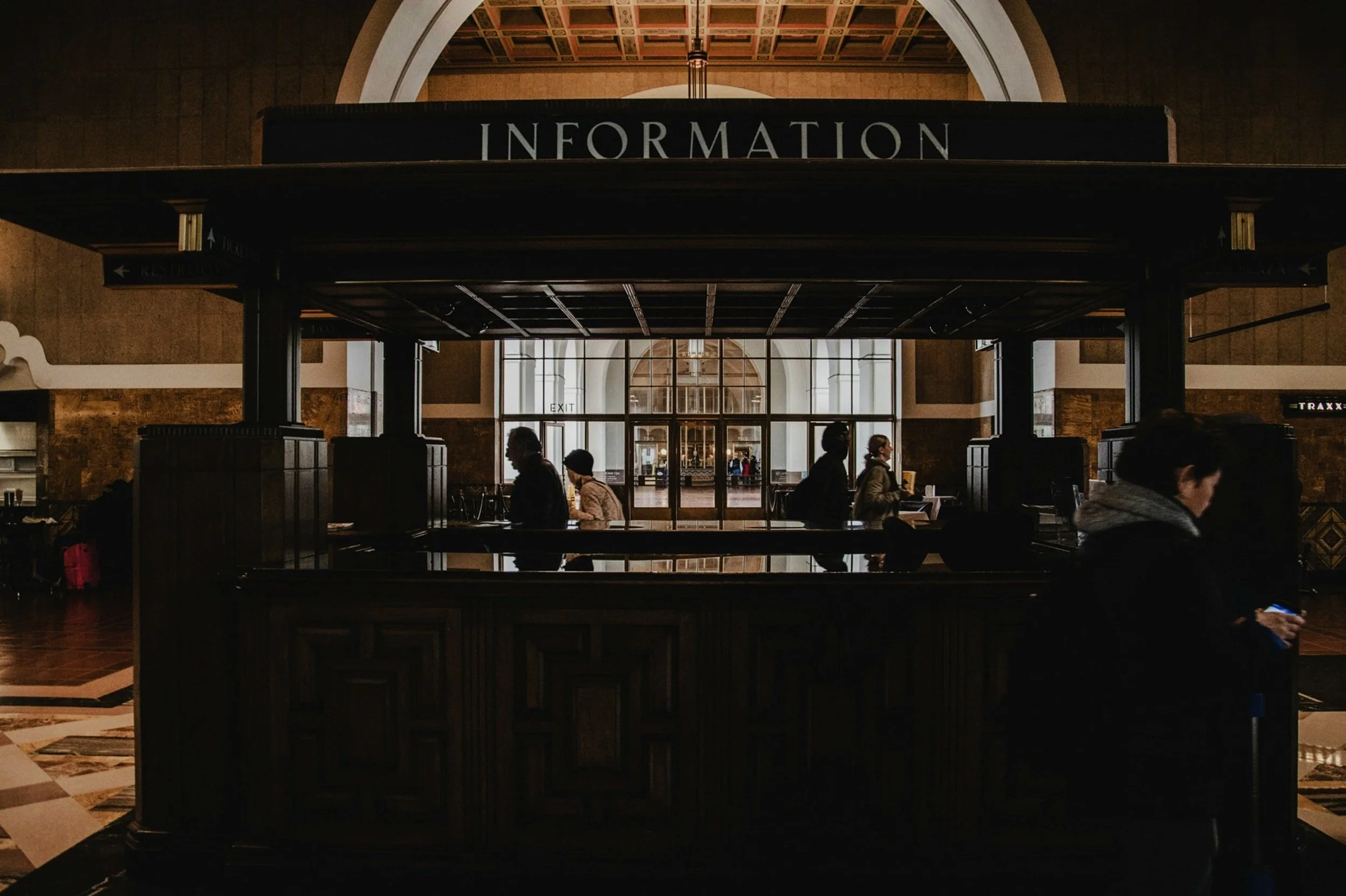 An airport or train station information booth with people walking in front. The sign reads 'INFORMATION' and is mounted above the booth. There is an arched ceiling and large windows in the background.