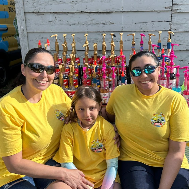Three people wearing yellow shirts sitting together outdoors with colorful soap bottles in the background, smiling for a photo.