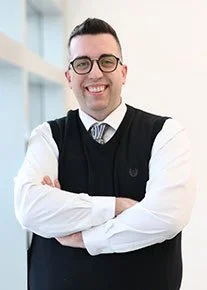 A smiling man wearing glasses and a light-colored blazer standing in front of yellow autumn leaves.