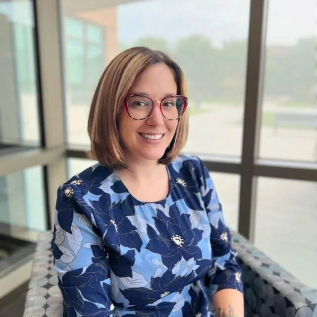 A woman with shoulder-length light brown hair wearing glasses and a blue floral blouse, sitting indoors near large windows.