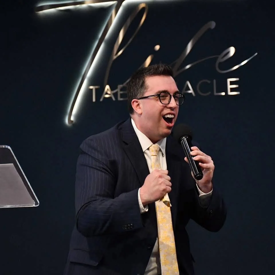A smiling man wearing glasses and a light-colored blazer standing in front of yellow autumn leaves.