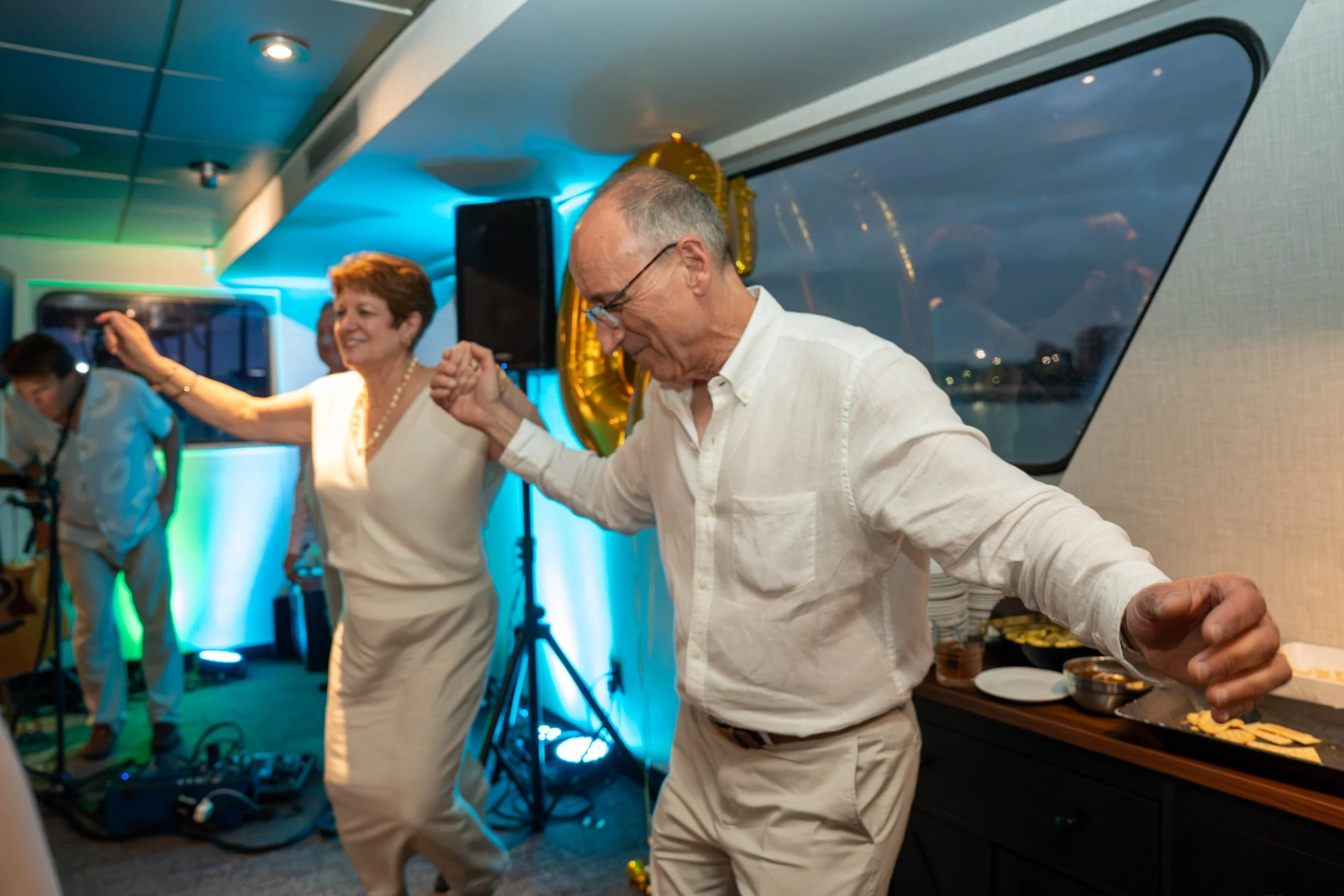 Older couple dancing at a brightly lit indoor party with a DJ, gold balloons, and a buffet table.