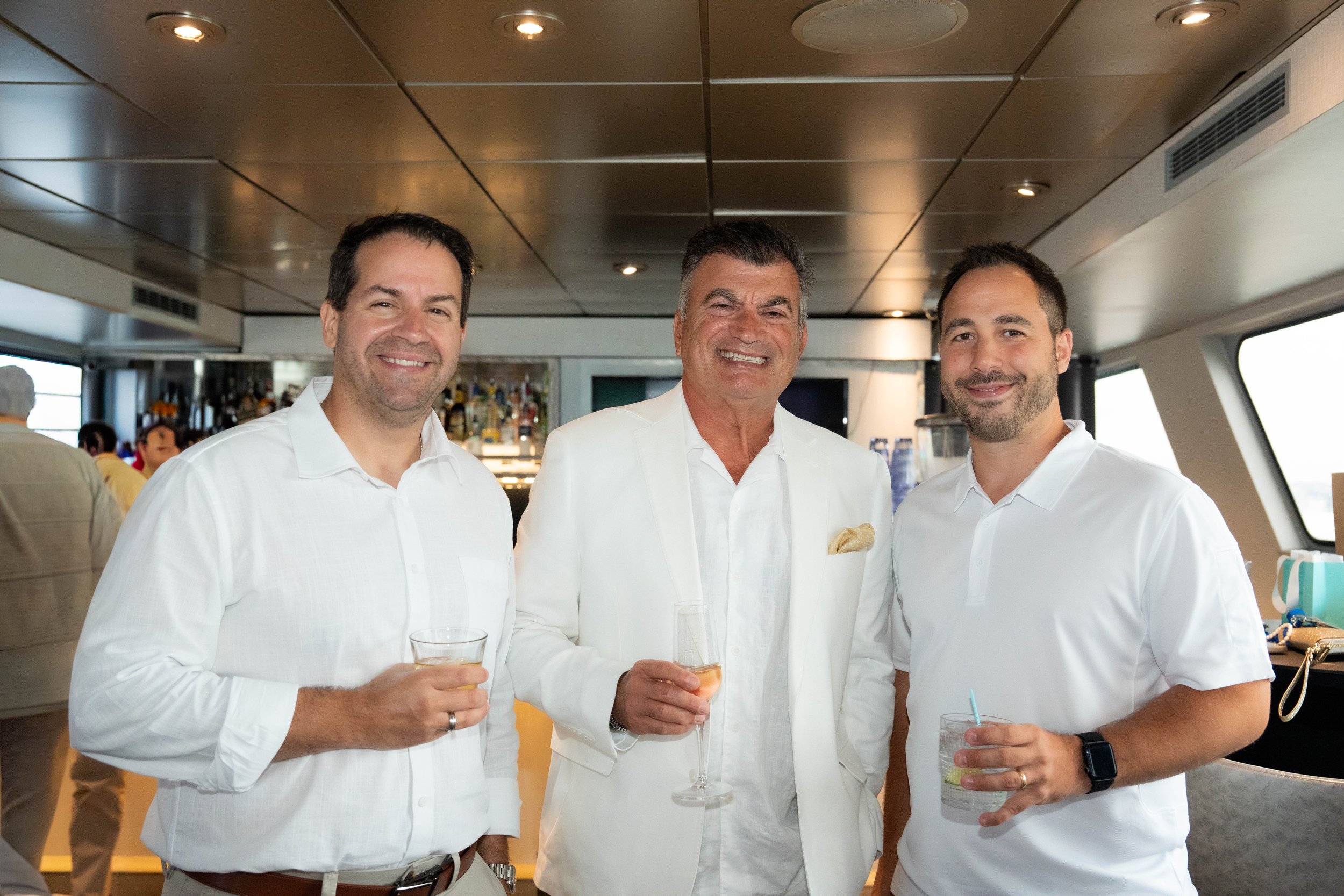 Three men in white shirts holding drinks inside a yacht lounge.