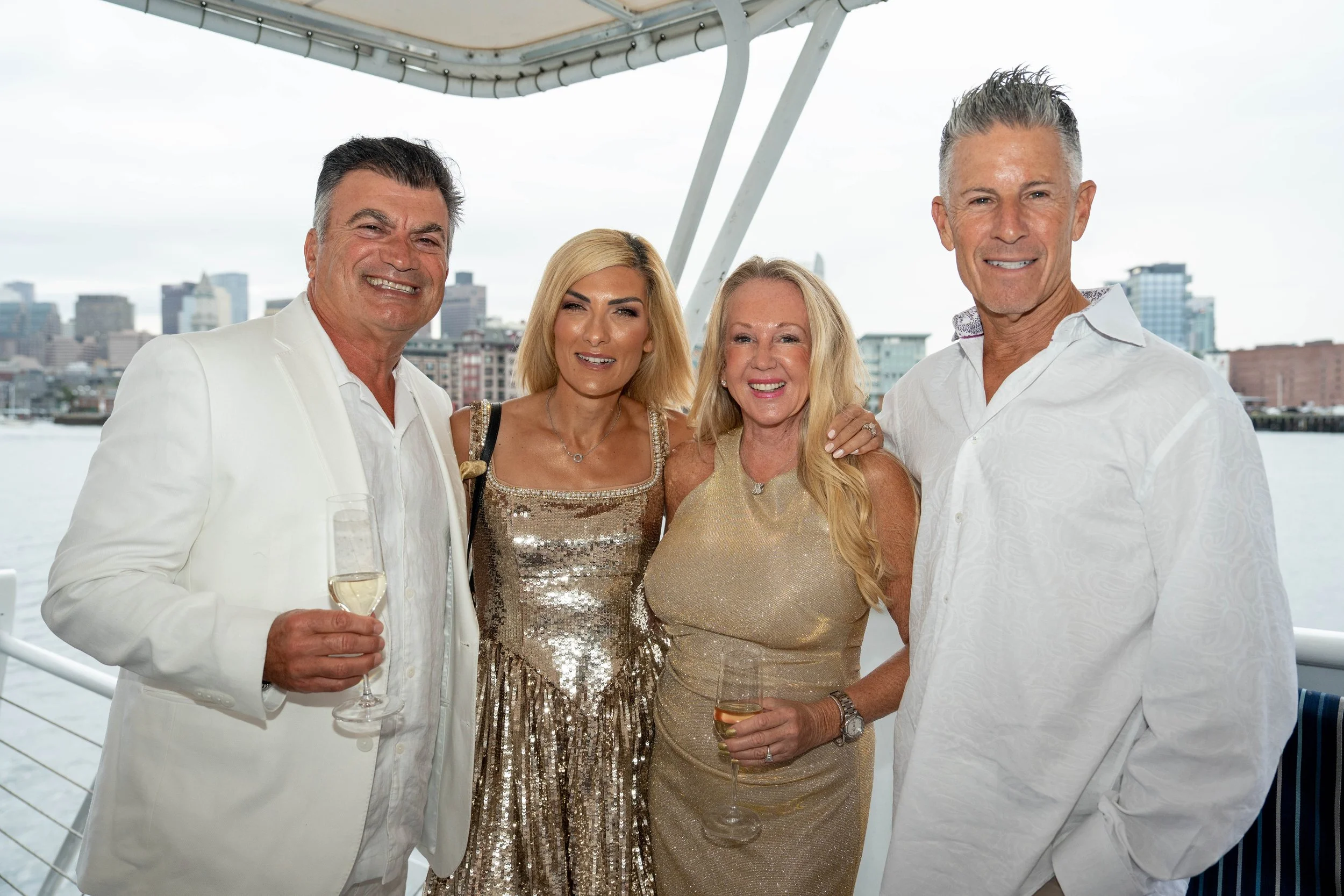 Four adults dressed in white and gold pose with drinks on a boat overlooking a city waterfront.