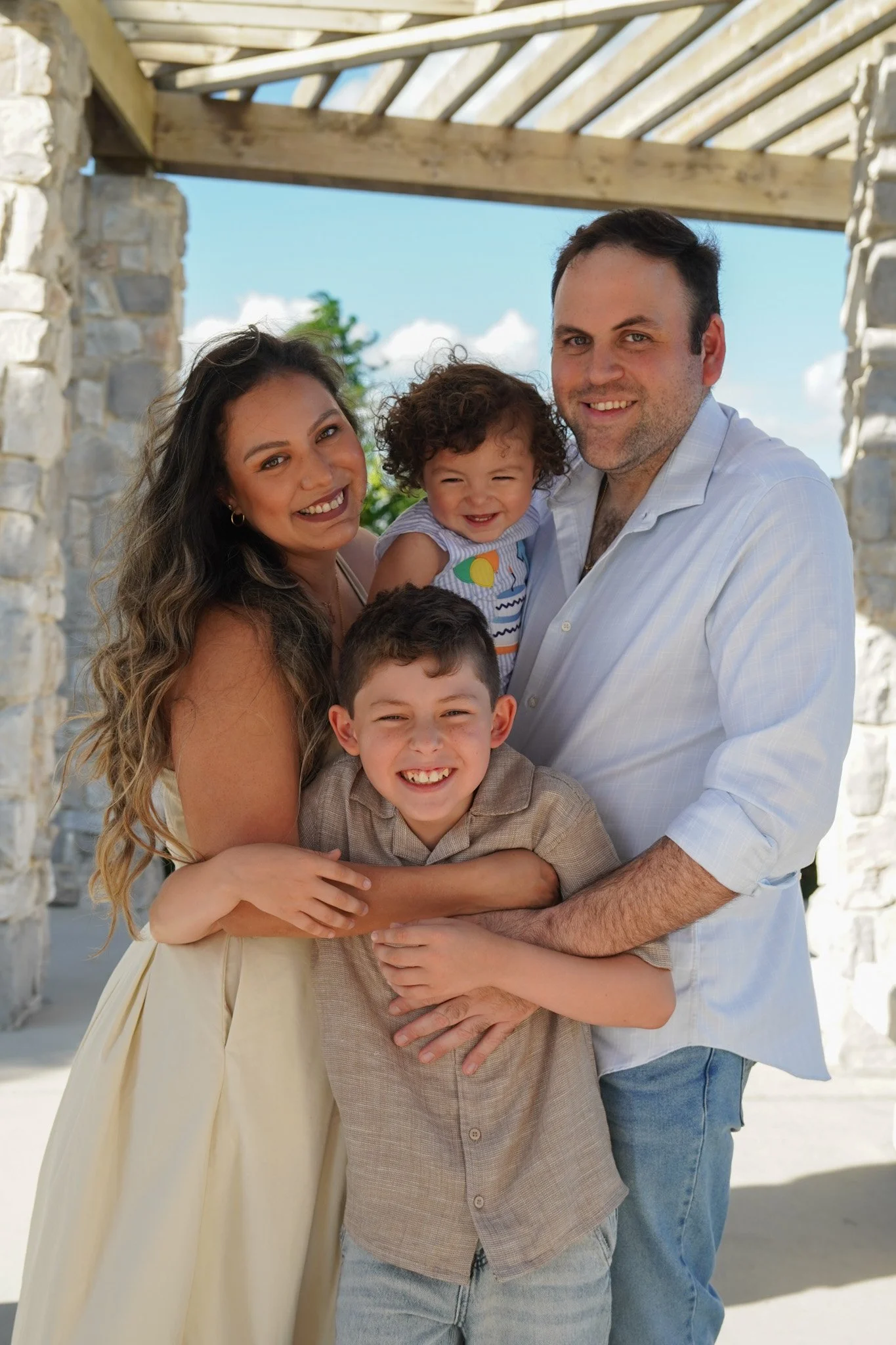 Smiling family of four posing together beneath a rustic stone pergola in bright sunlight